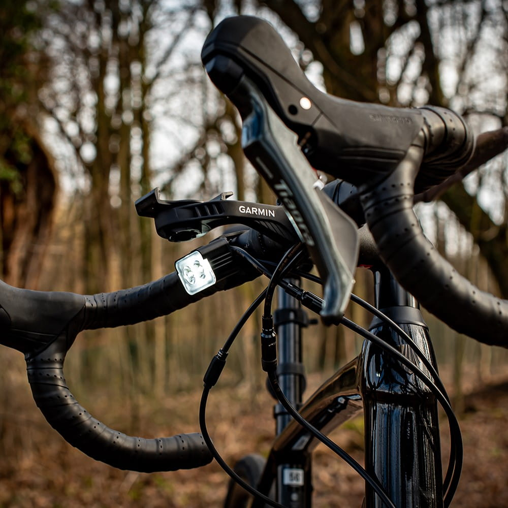 Czarna kierownica roweru z komputerem rowerowym Garmin i światłem. Tło to rozmyty las.