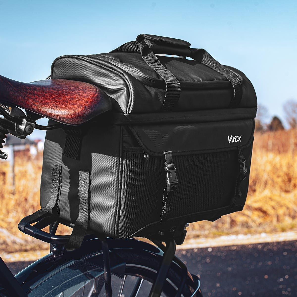 Czarna torba rowerowa zamontowana na rowerze. Widoczne brązowe siodełko. Tło to słoneczna sceneria zewnętrzna.