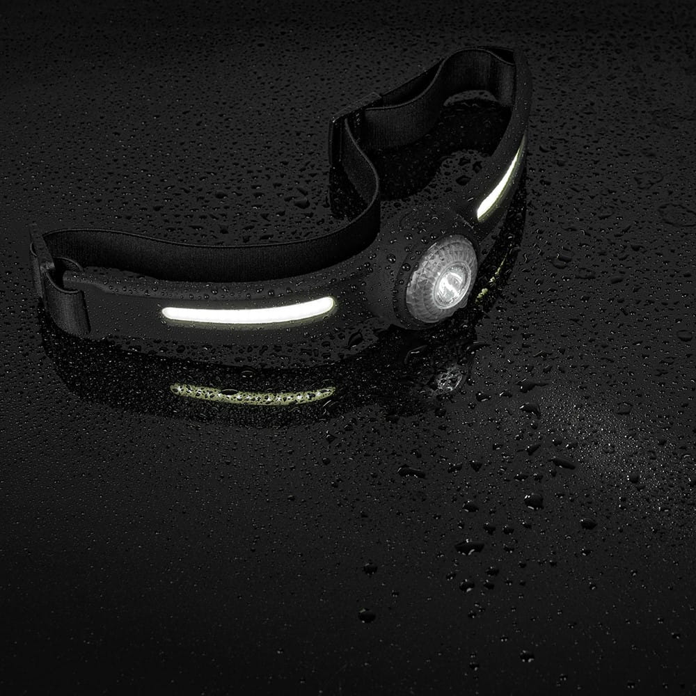 Czarna lampa czołowa na mokrej, czarnej powierzchni. Emitowane białe światło. Widoczne krople wody.