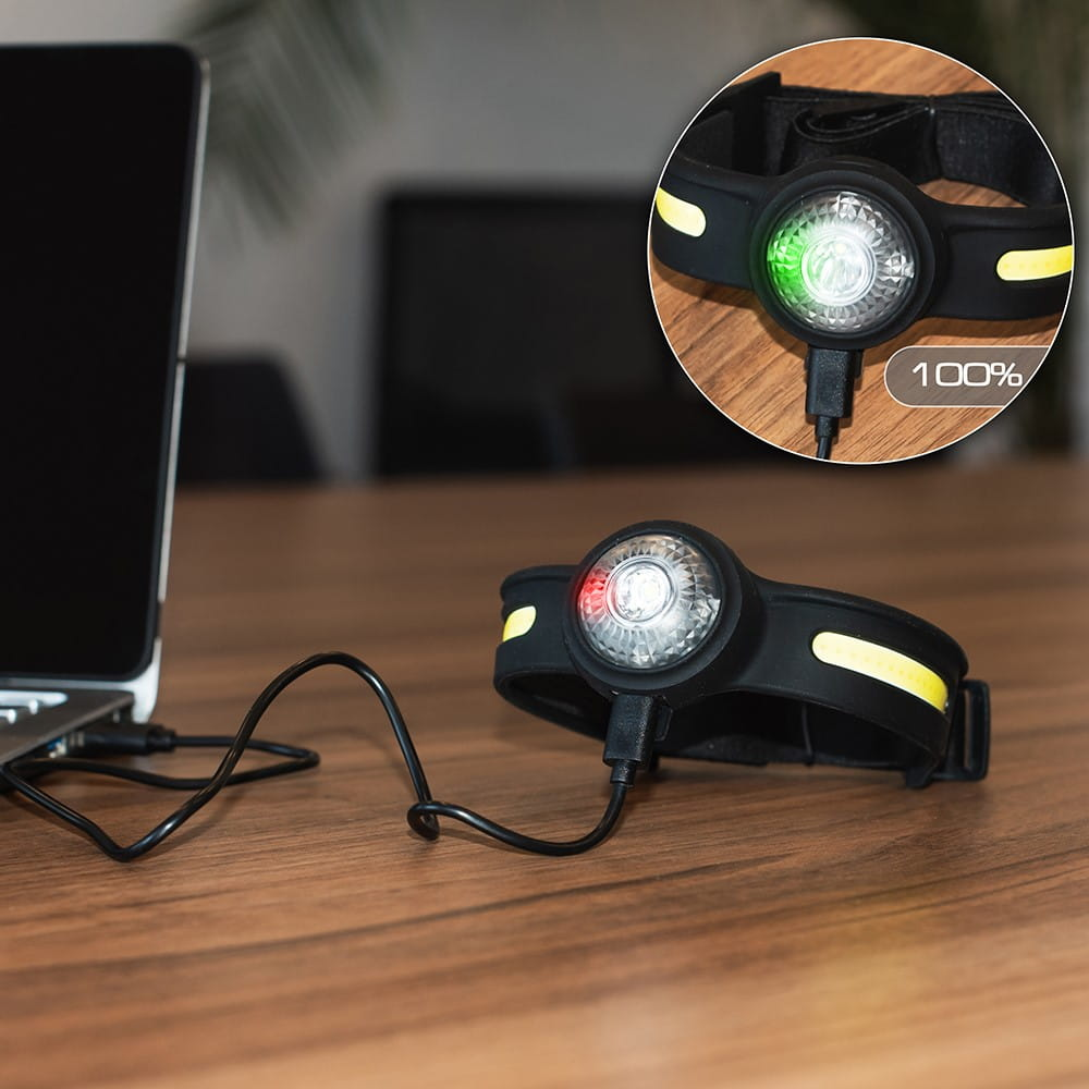 Lampa czołowa ładująca się na drewnianym stole. Wyświetlacz pokazuje 100% naładowania. Druga lampa czołowa obok.