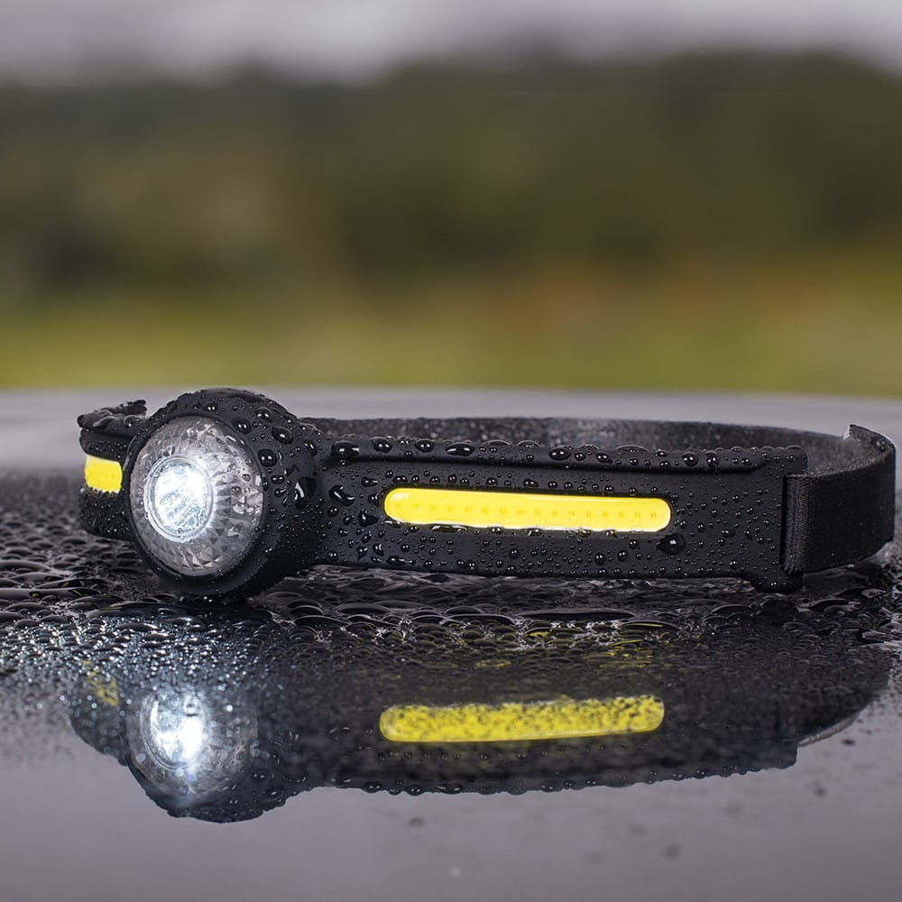 Czarna lampa czołowa z białym światłem na mokrej powierzchni. Żółte akcenty na pasku. Zielone tło.