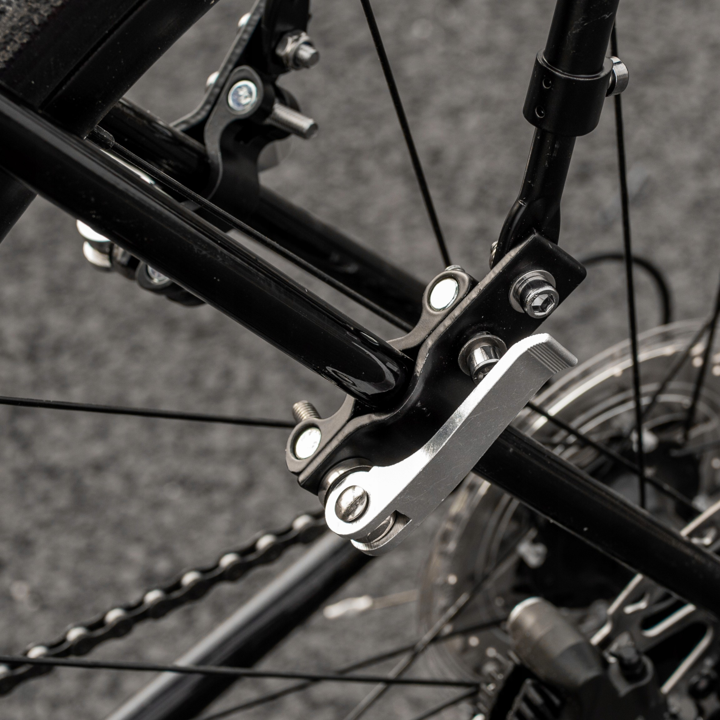 Zbliżenie na tylne koło roweru. Widoczna czarna rama roweru, łańcuch i srebrna dźwignia.