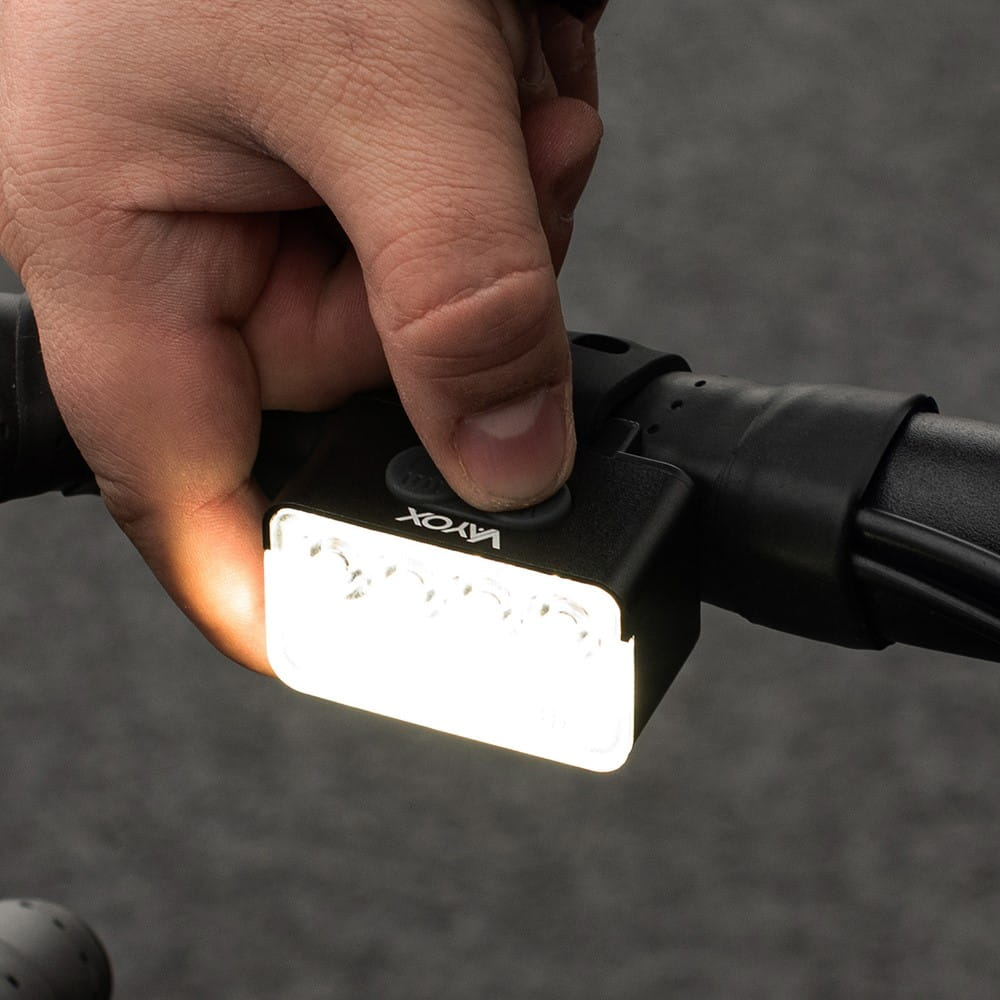 Czarna lampa rowerowa Vayox przymocowana do kierownicy, podświetlona. Ręka trzyma ją.