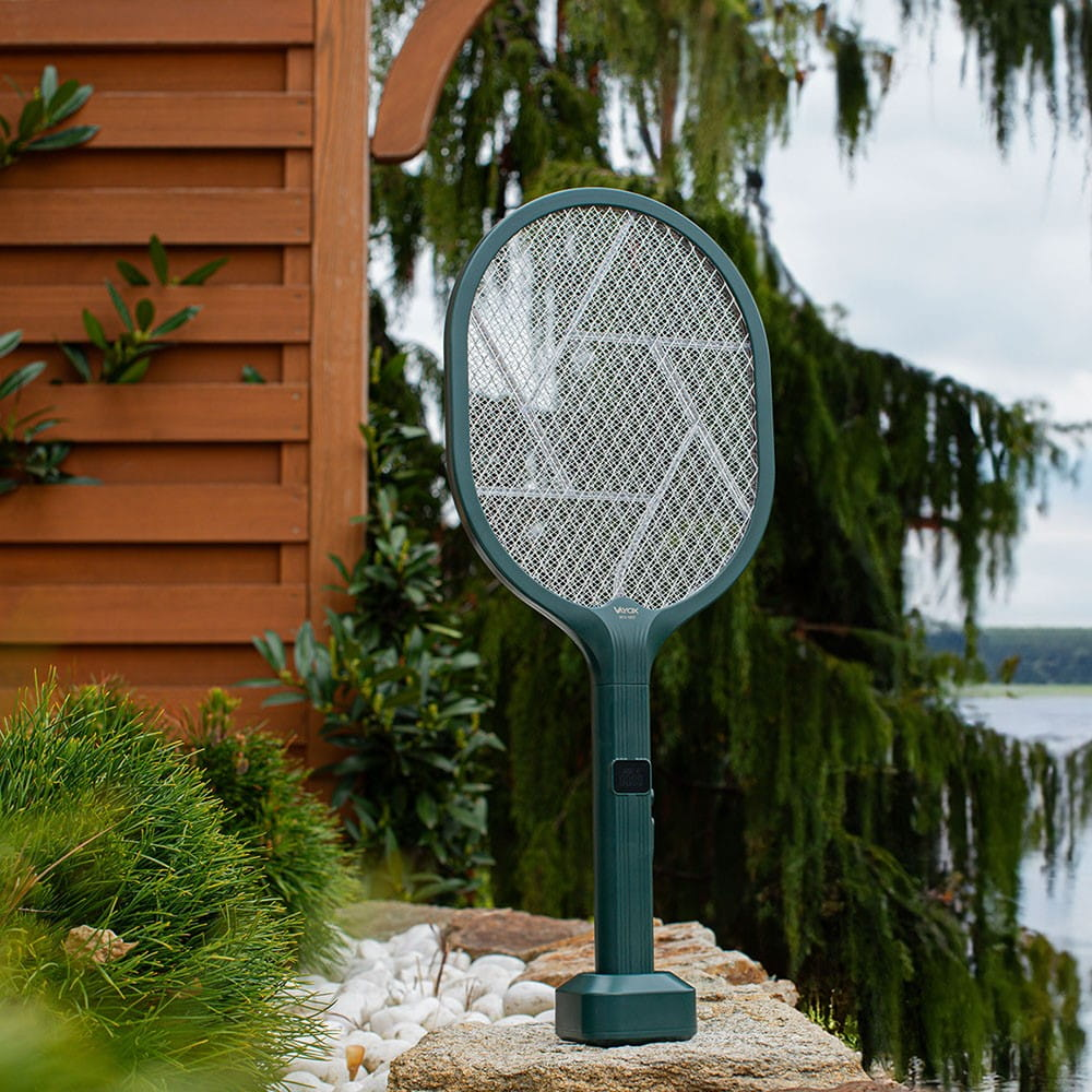 Zielona lampa owadobójcza na skale, przed liśćmi i jeziorem. Pochmurne niebo.