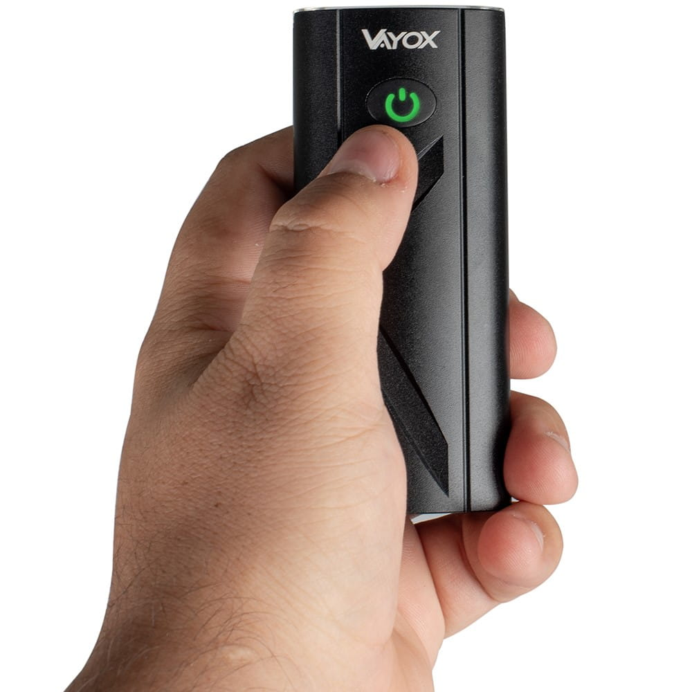 Ręka trzymająca czarne urządzenie Vayox z zielonym przyciskiem zasilania.