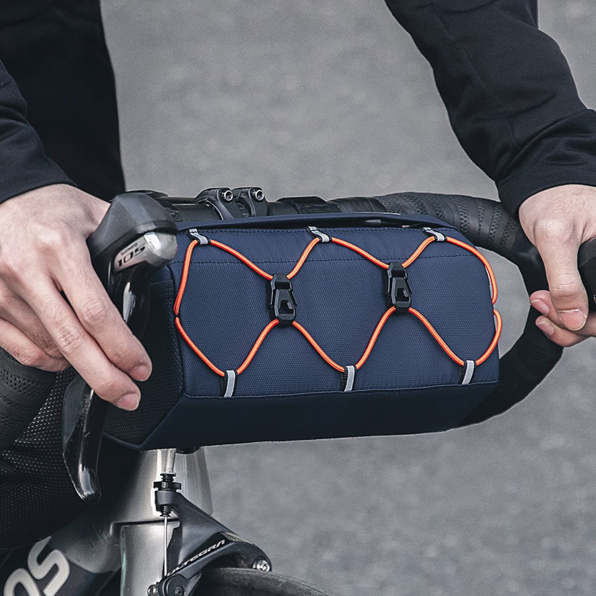 Kolarz trzyma niebieską torbę na kierownicę z pomarańczowymi elastycznymi sznurkami na kierownicy roweru.
