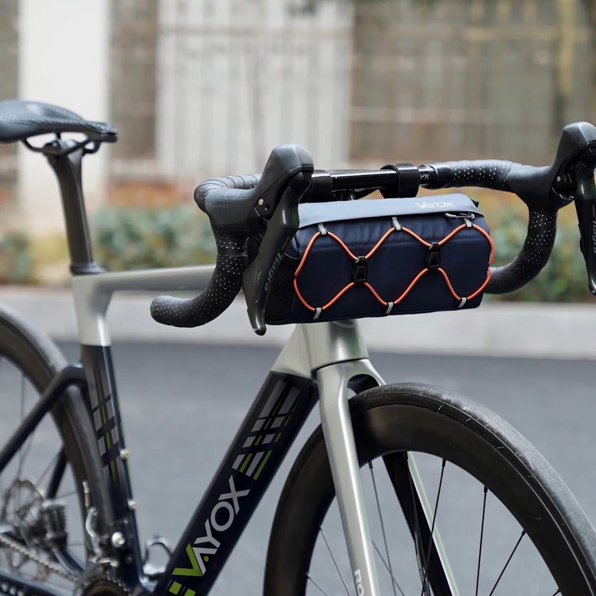 Kierownica roweru z przymocowaną granatową torbą, pomarańczowe paski. Scena uliczna.