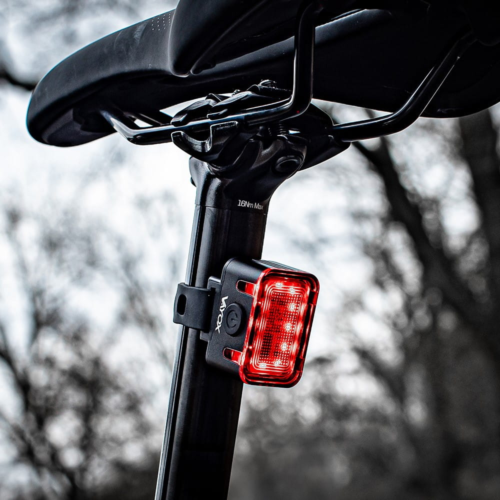 Czerwone tylne światło rowerowe marki VOX jest przymocowane do czarnej sztycy podsiodłowej. Tło jest rozmyte.
