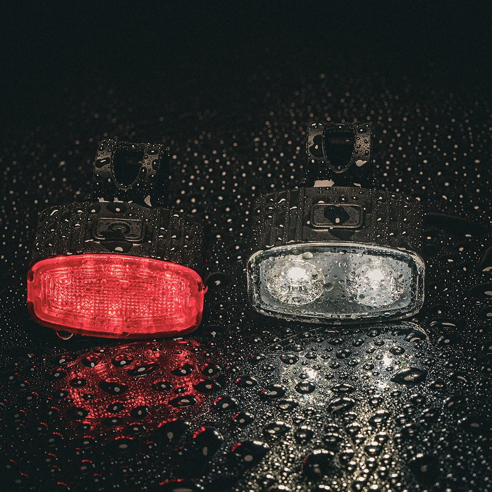 Dwa światła rowerowe, jedno czerwone, jedno białe, z kroplami wody na ciemnej powierzchni.