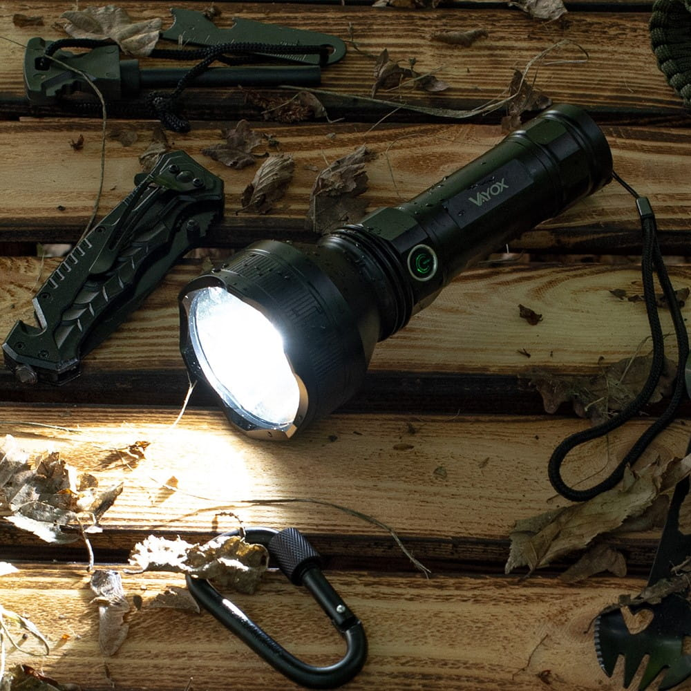 Czarna latarka na drewnie, z nożem i innymi narzędziami. Latarka emituje jasne światło.