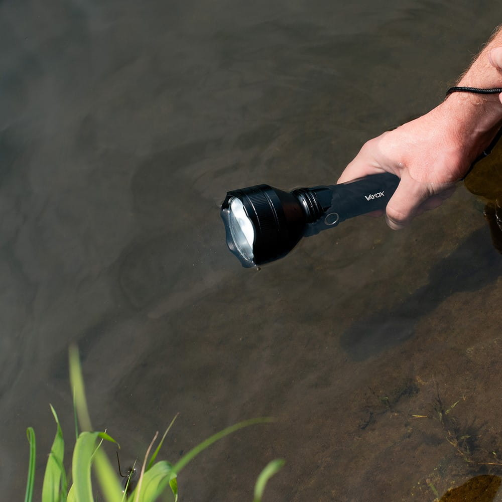 Ręka trzymająca czarną latarkę nad wodą. Latarka jest włączona i świeci w wodzie.