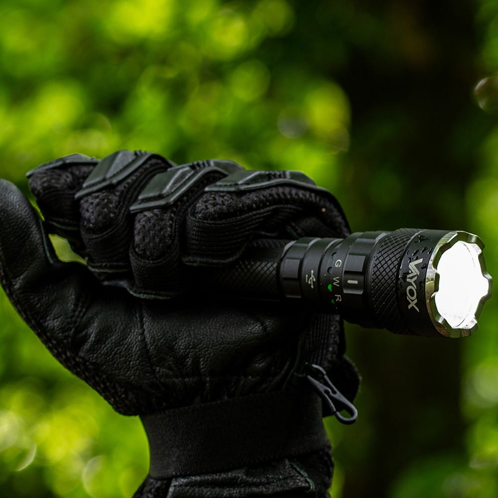 Ręka w rękawicy trzymająca latarkę VAYOX, skierowana w stronę kamery, rozmyte tło.