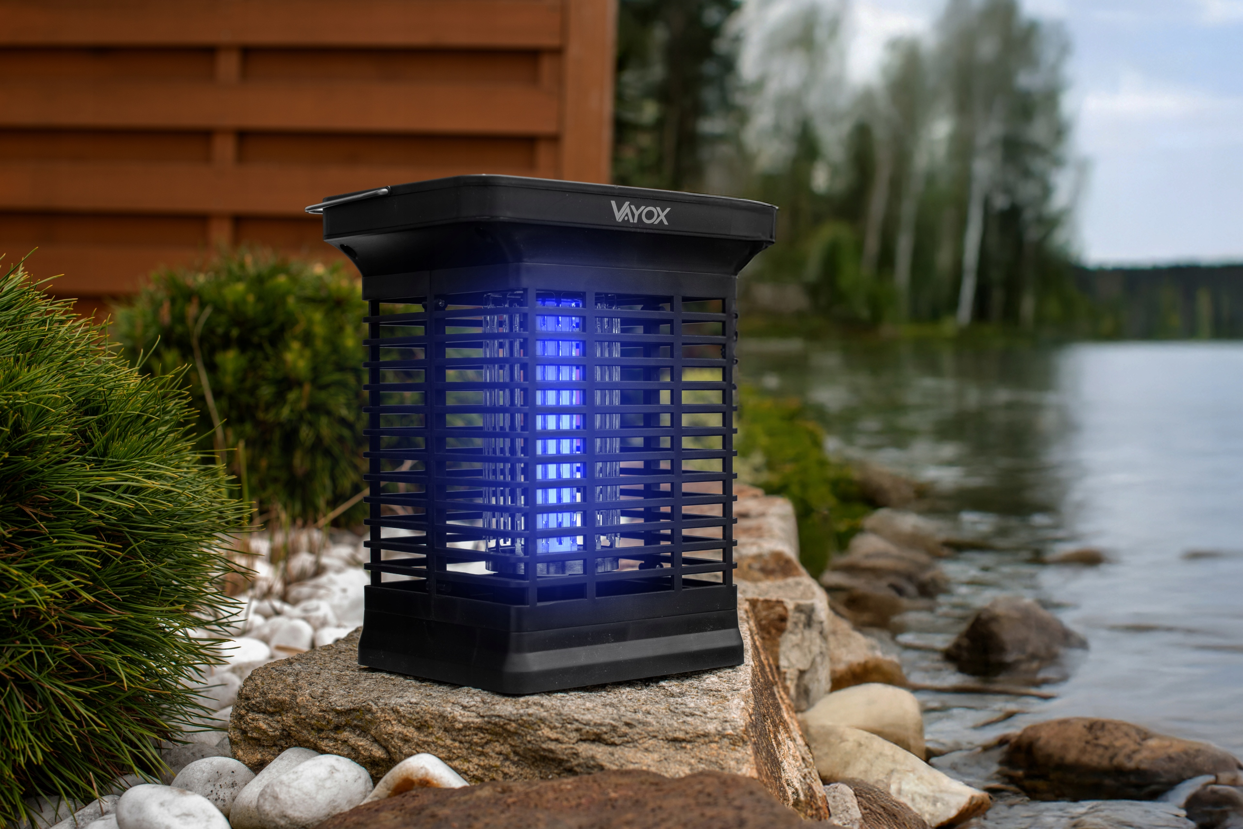 Czarny zapper na owady z niebieskim światłem, ustawiony na zewnątrz w pobliżu wody i zieleni.