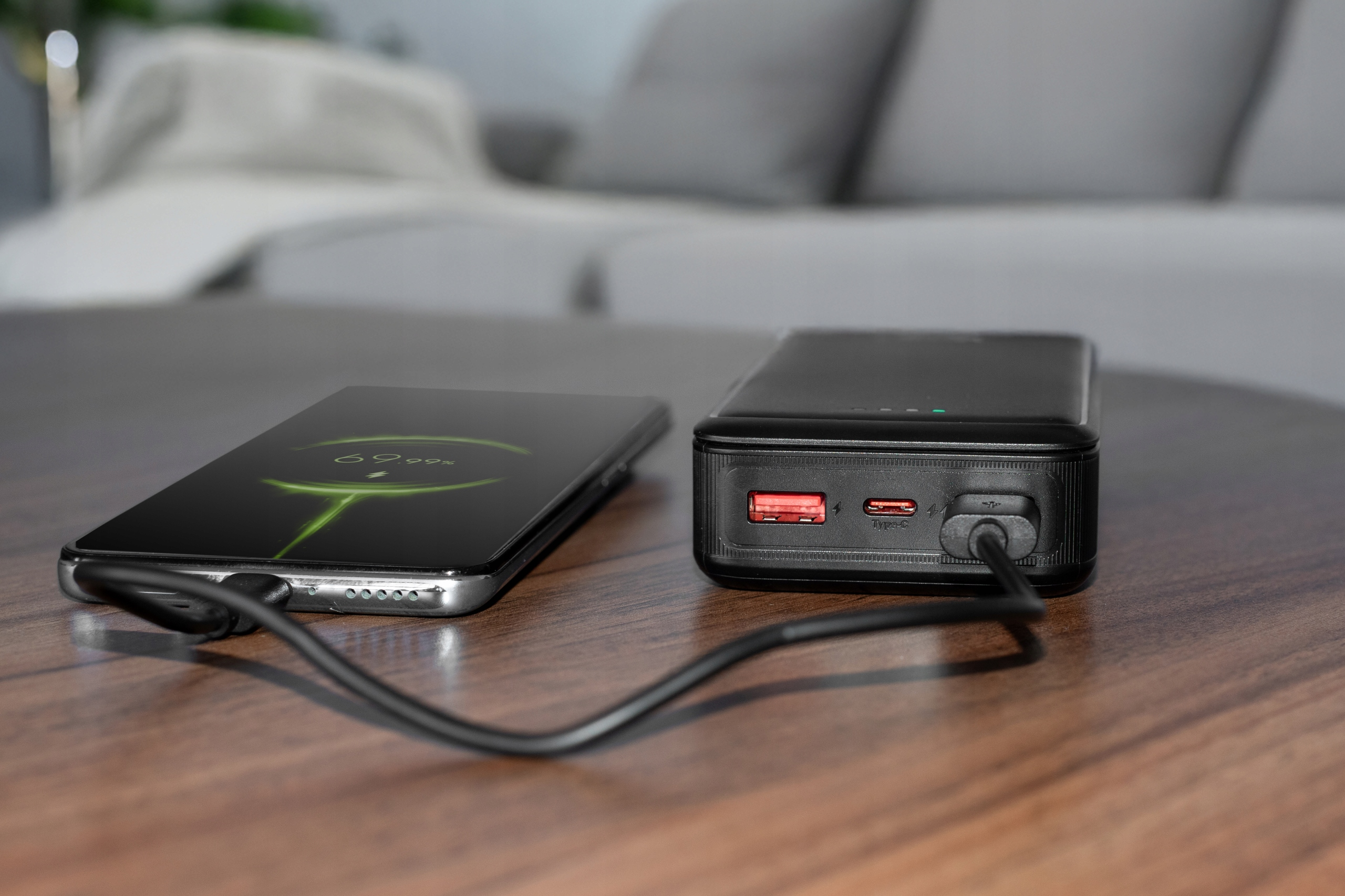 Telefon ładujący się na czarnym power banku. Telefon pokazuje procent naładowania. Drewniany stół.