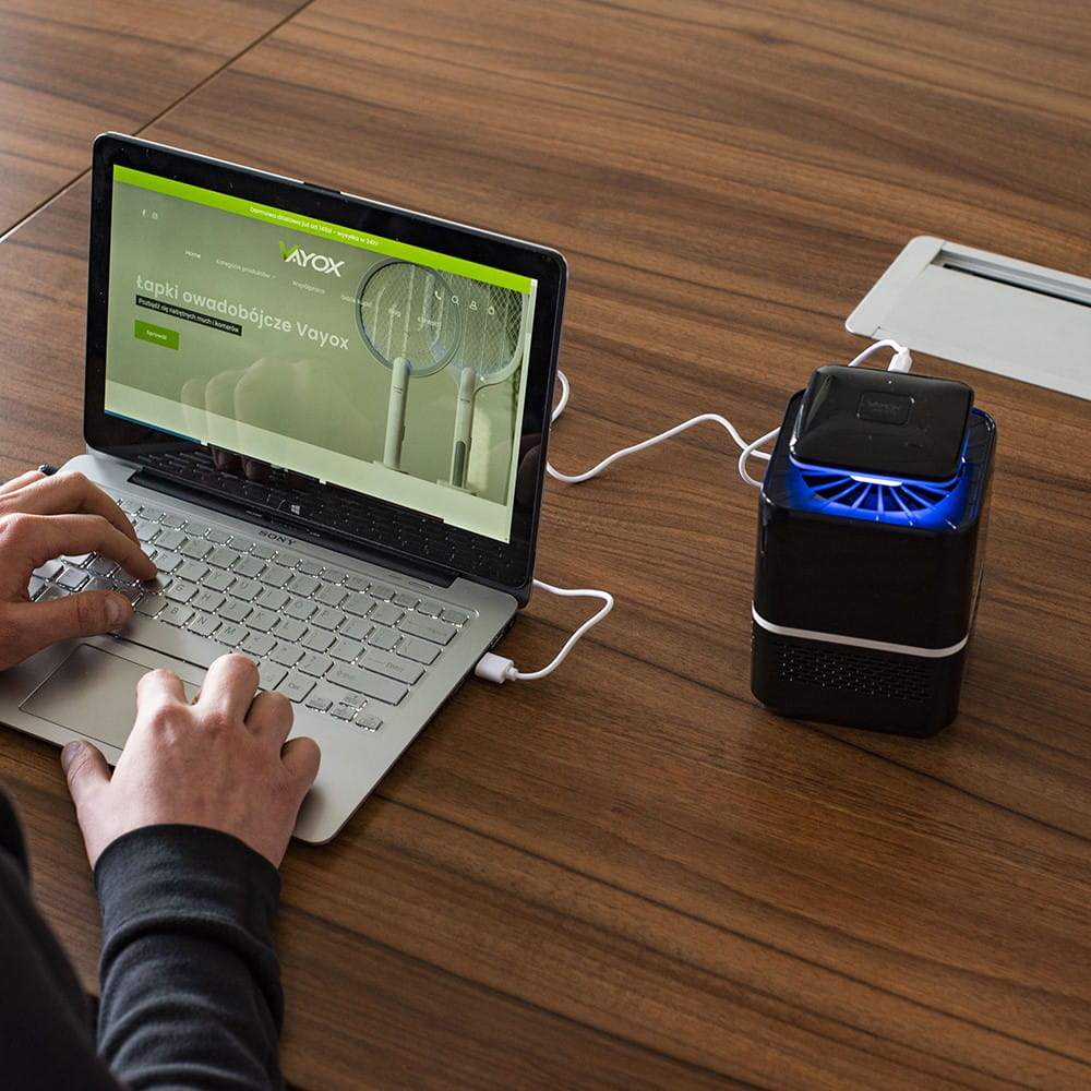 Osoba korzysta z laptopa, obok czarnego łapacza owadów. Łapacz jest podłączony do laptopa białym kablem.