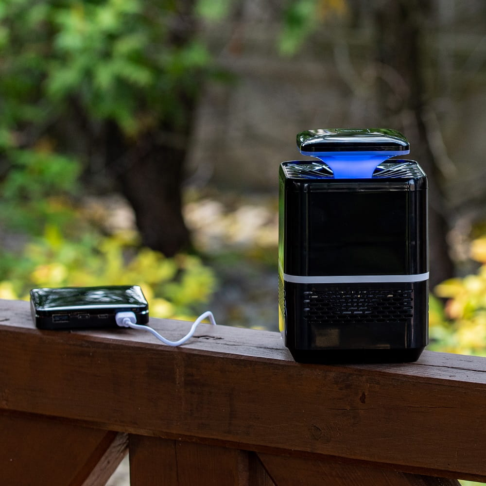 Czarny łapacz owadów z niebieskim światłem na drewnianej balustradzie. Power bank jest podłączony białym kablem.