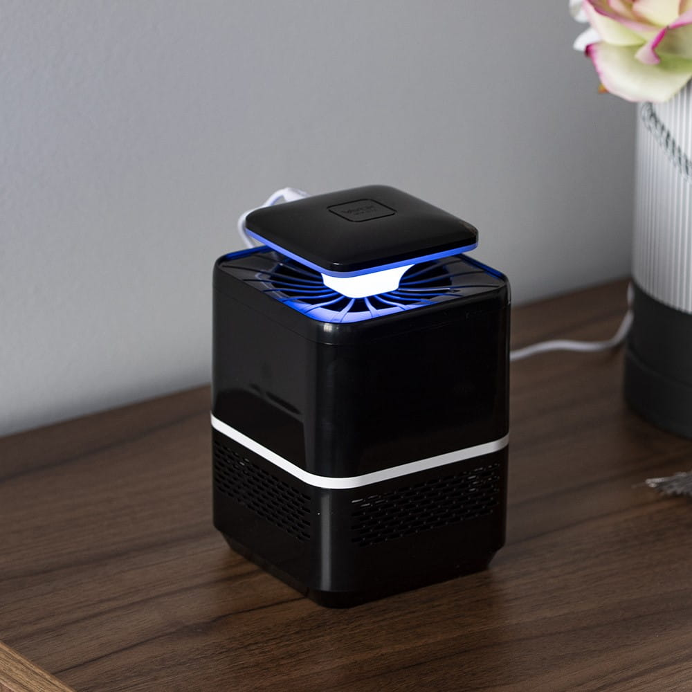 Czarny łapacz owadów z włączonym niebieskim światłem. Łapacz stoi na drewnianym stole z kwiatem w tle.