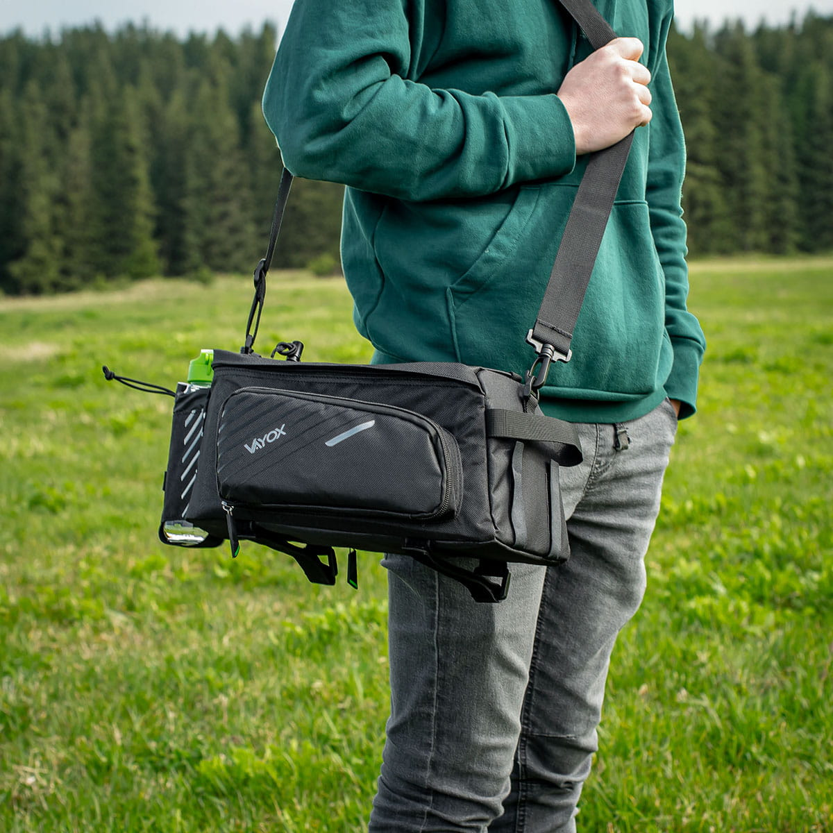 Osoba z czarną torbą na ramieniu, idąca po zielonej trawie.