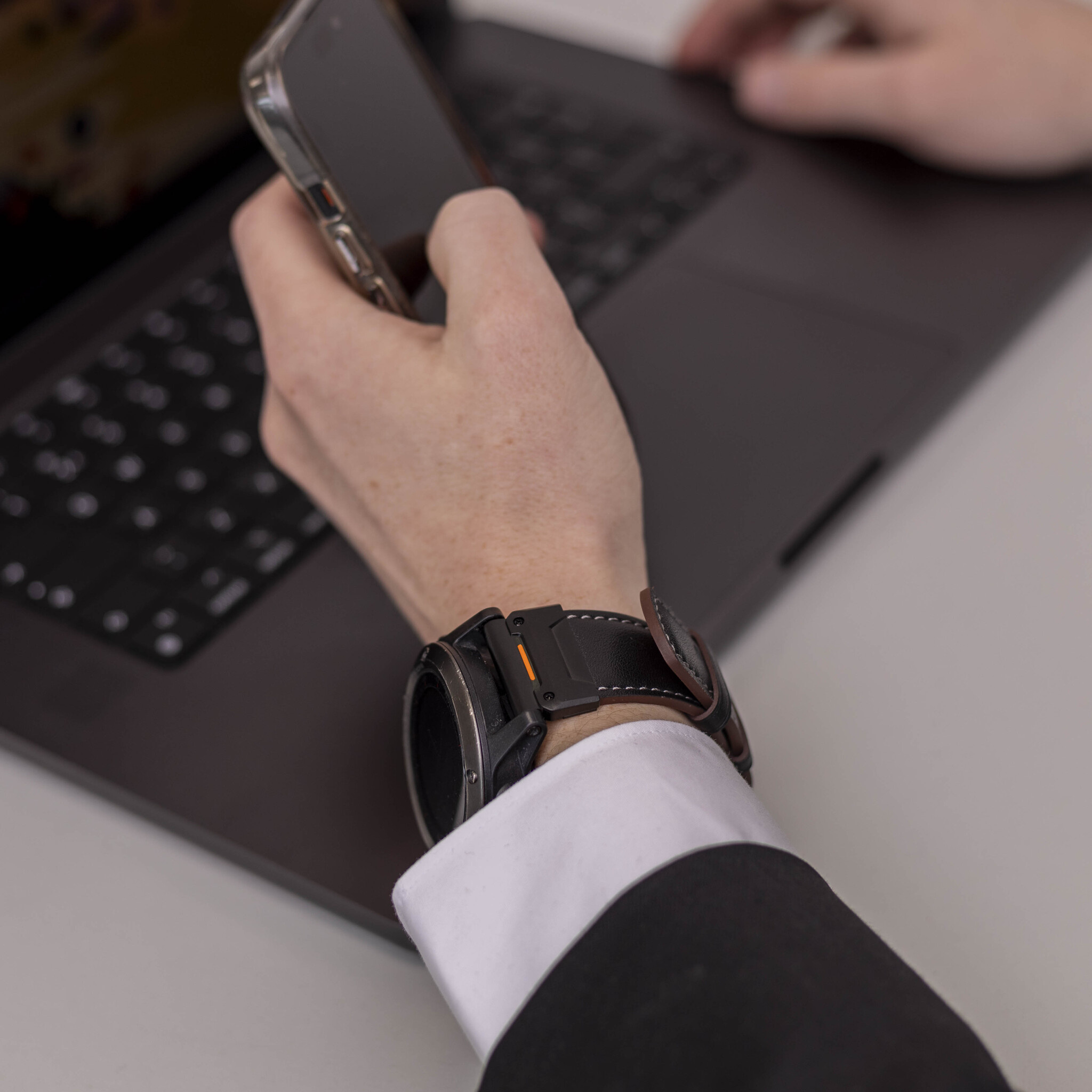 Eine Person im Anzug hält ein Telefon in der Nähe eines Laptops und trägt eine schwarze Smartwatch.