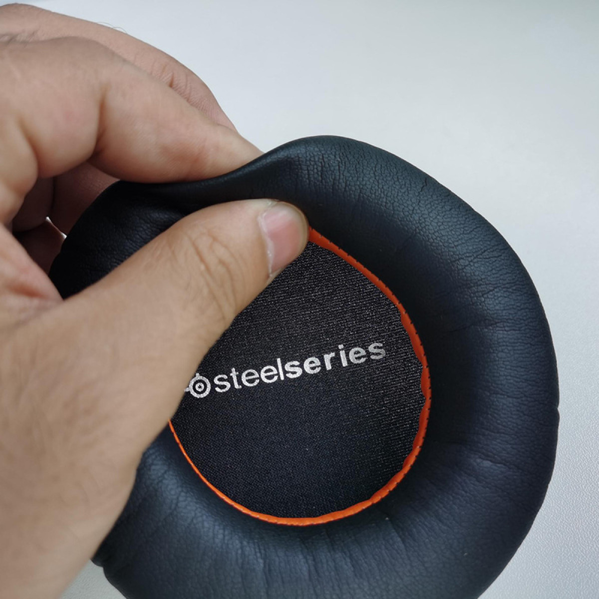 Eine Hand, die ein schwarzes Kopfhörer-Ohrpolster mit der Aufschrift 'steelseries' hält.