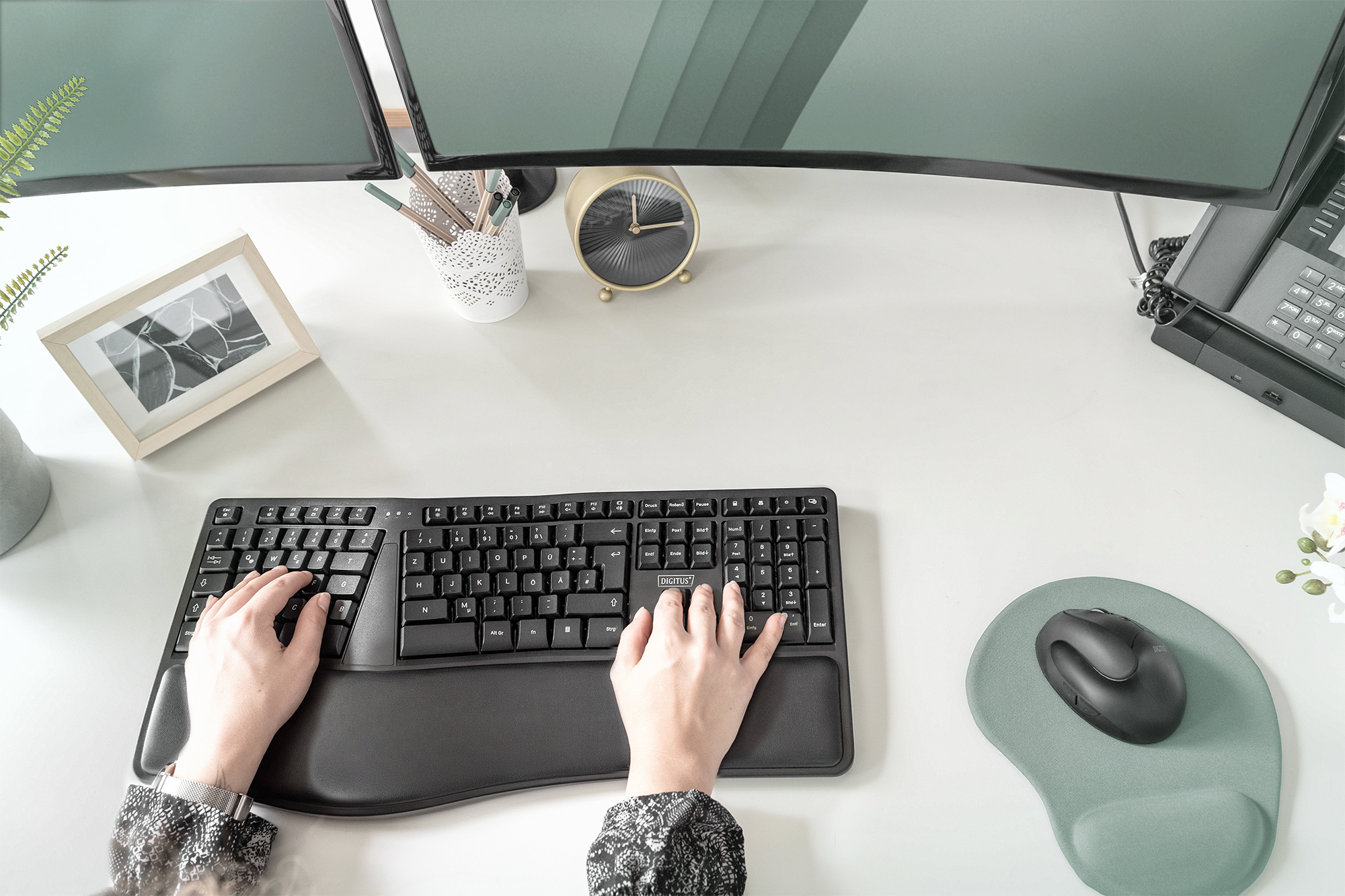 Mani che digitano su una tastiera ergonomica nera su una scrivania bianca. Sono presenti anche mouse e monitor.