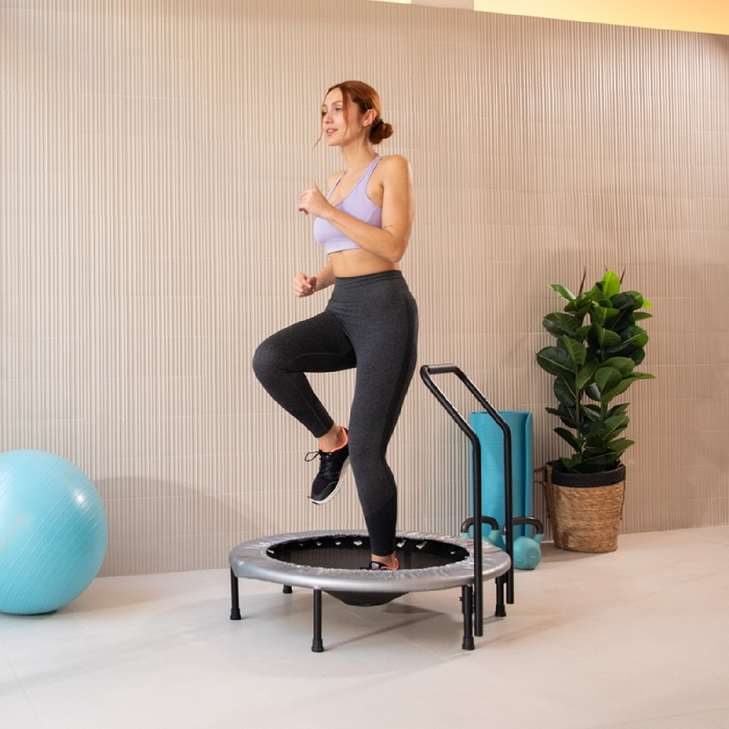 Kobieta ćwiczy na mini trampolinie. Tło: piłka do jogi, rośliny i ściana.