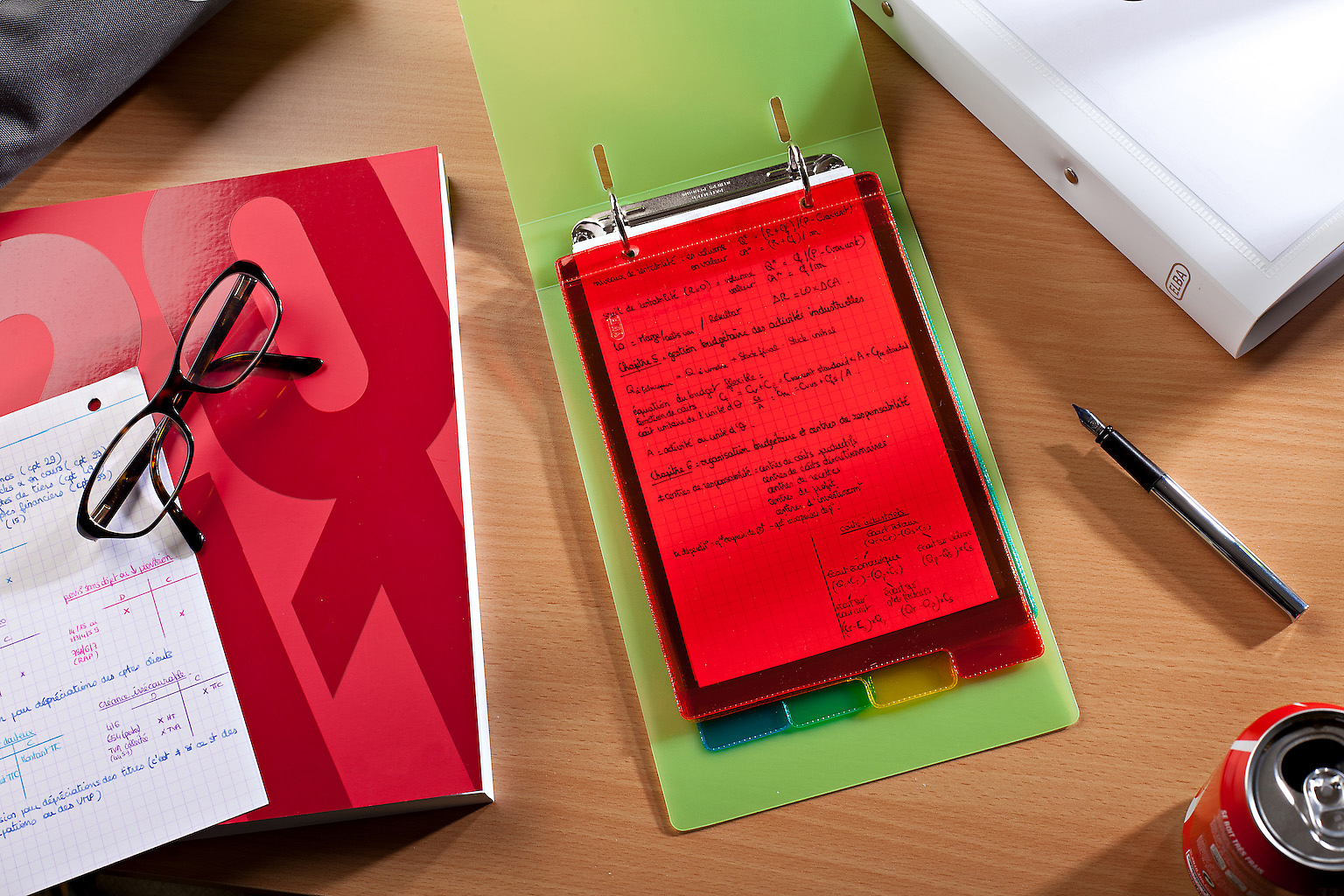 Scena na biurku: czerwony notatnik, okulary, notatki w teczce, długopis i puszka napoju gazowanego.