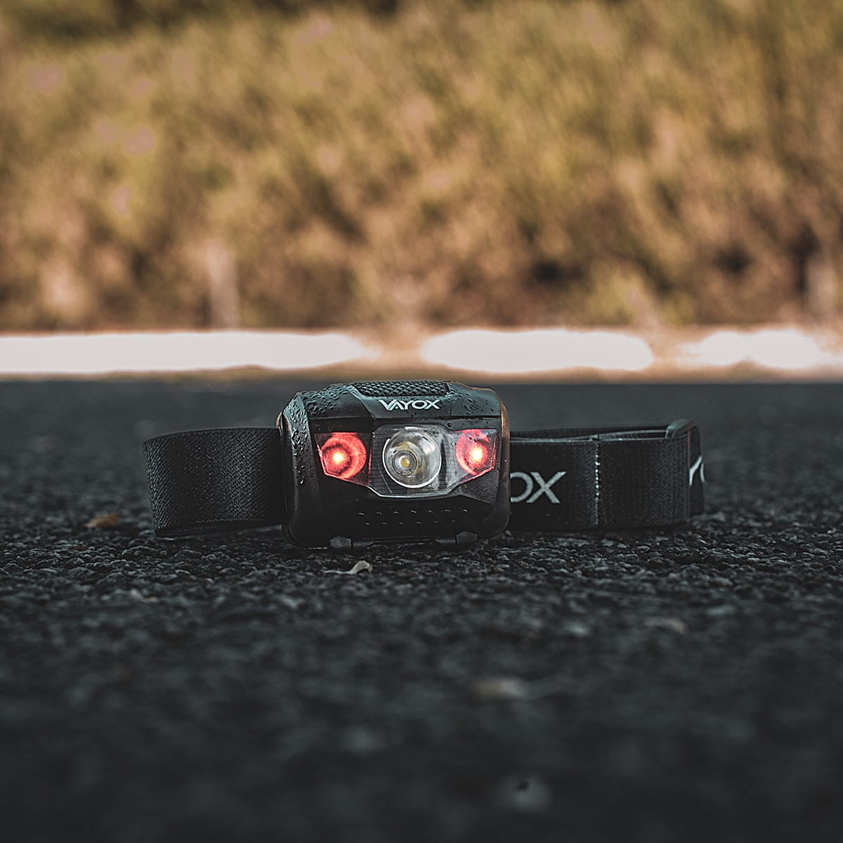 Czarna latarka czołowa z czerwonymi światłami bocznymi na ciemnej nawierzchni asfaltowej.