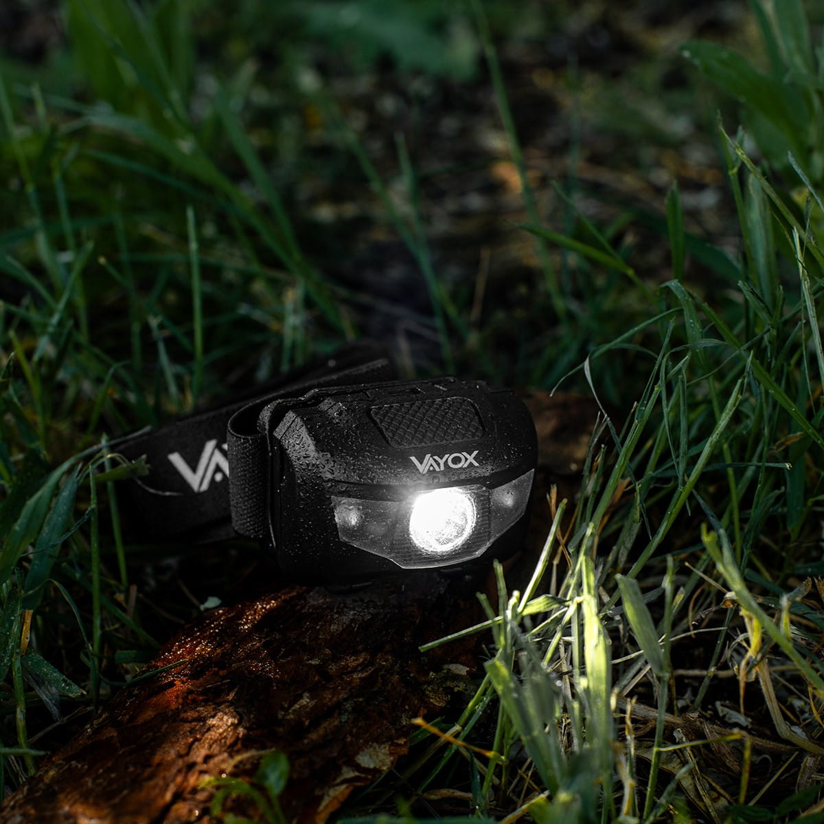 Czarna latarka czołowa na kłodzie pokrytej zieloną trawą. Latarka czołowa ma na sobie markę 'VAYOX'.
