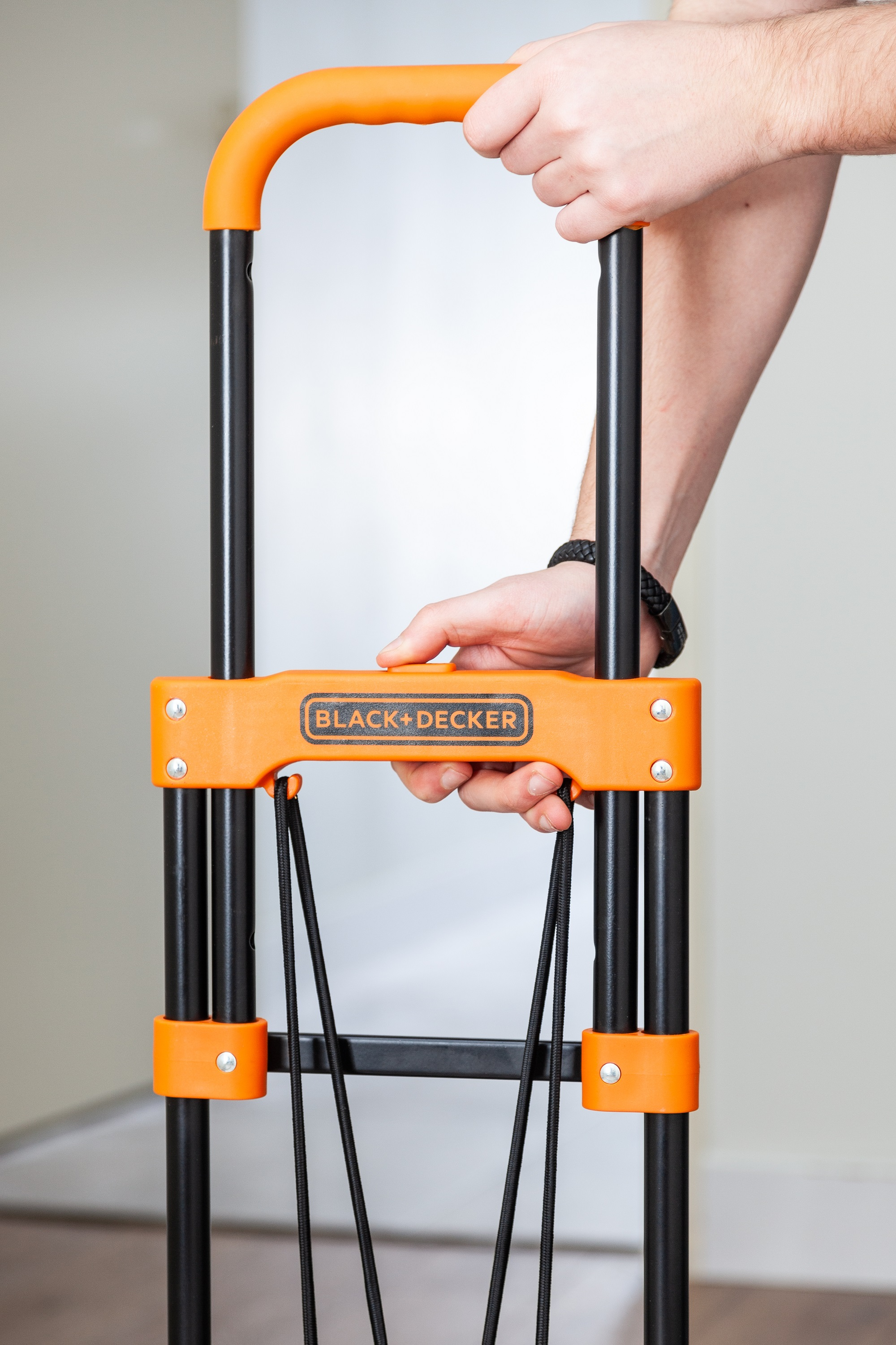 Close-up van de handen van een persoon die een zwarte en oranje opvouwbare handtruck vasthoudt.