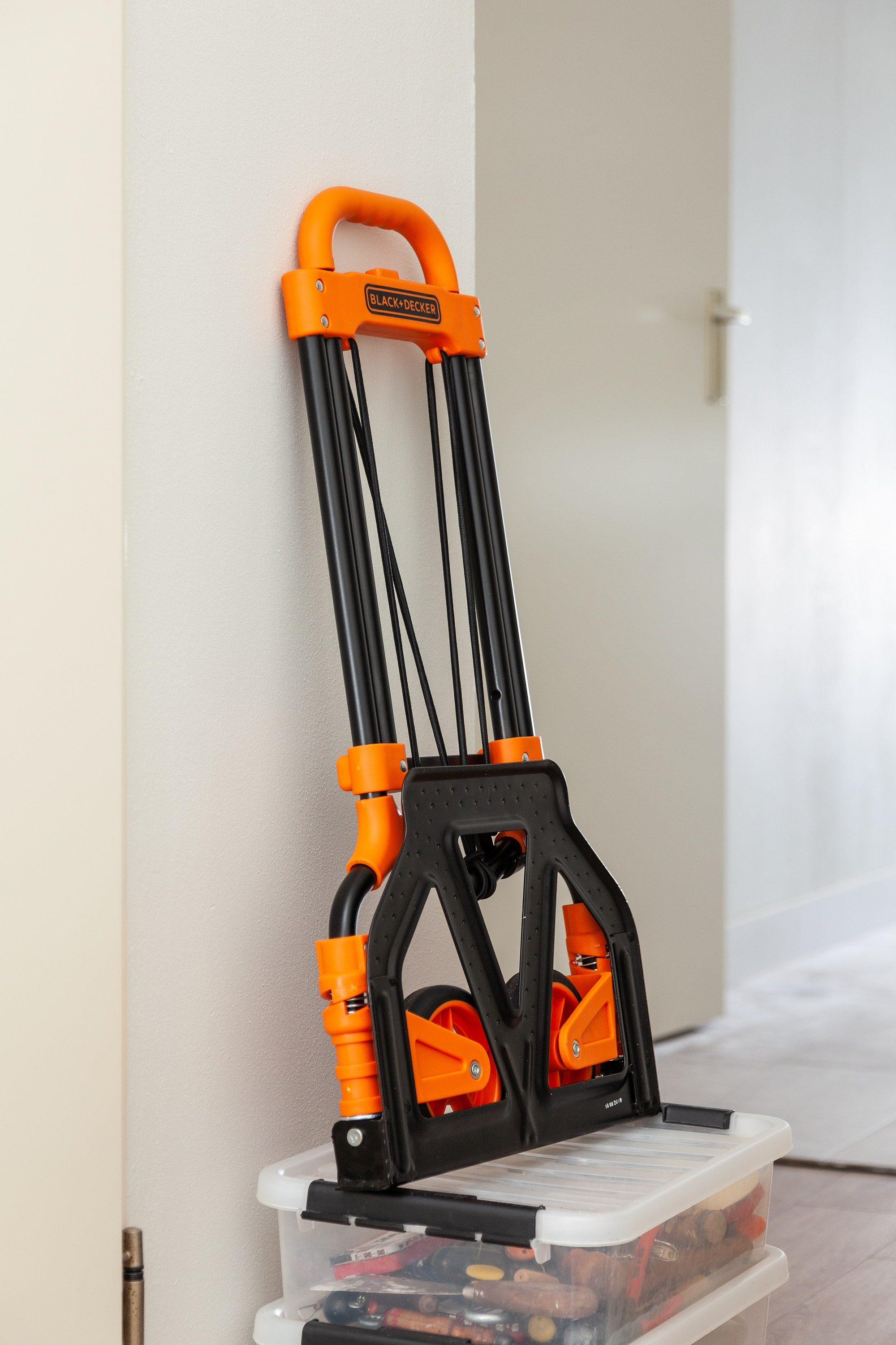 Een oranje en zwarte handtruck die tegen een muur leunt naast een doorzichtige opbergcontainer.