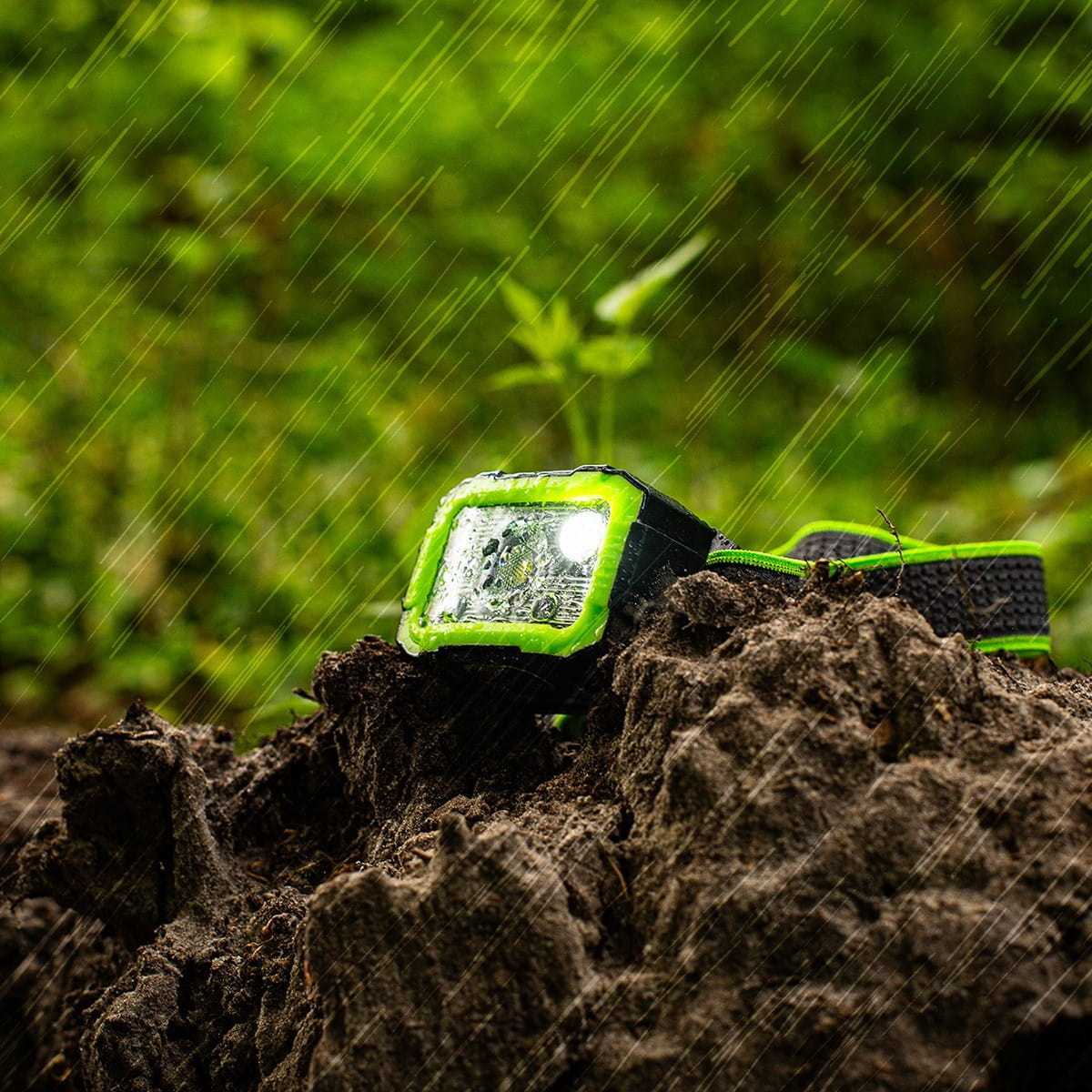 Latarka czołowa na błotnistej powierzchni z deszczem. Zielona i czarna, światło włączone.