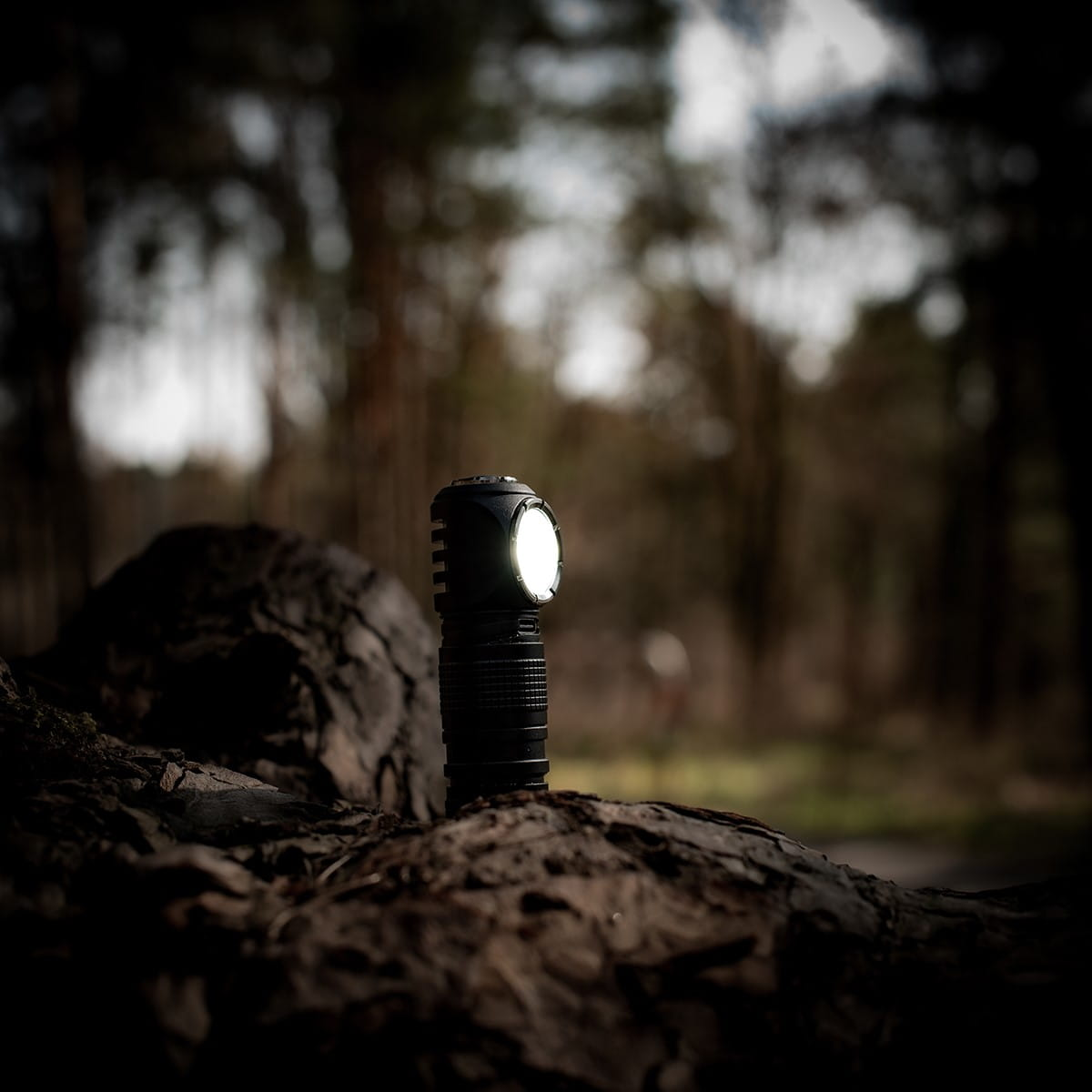 Czarna latarka czołowa z włączonym światłem, na rozmytym tle drzew i lasu.
