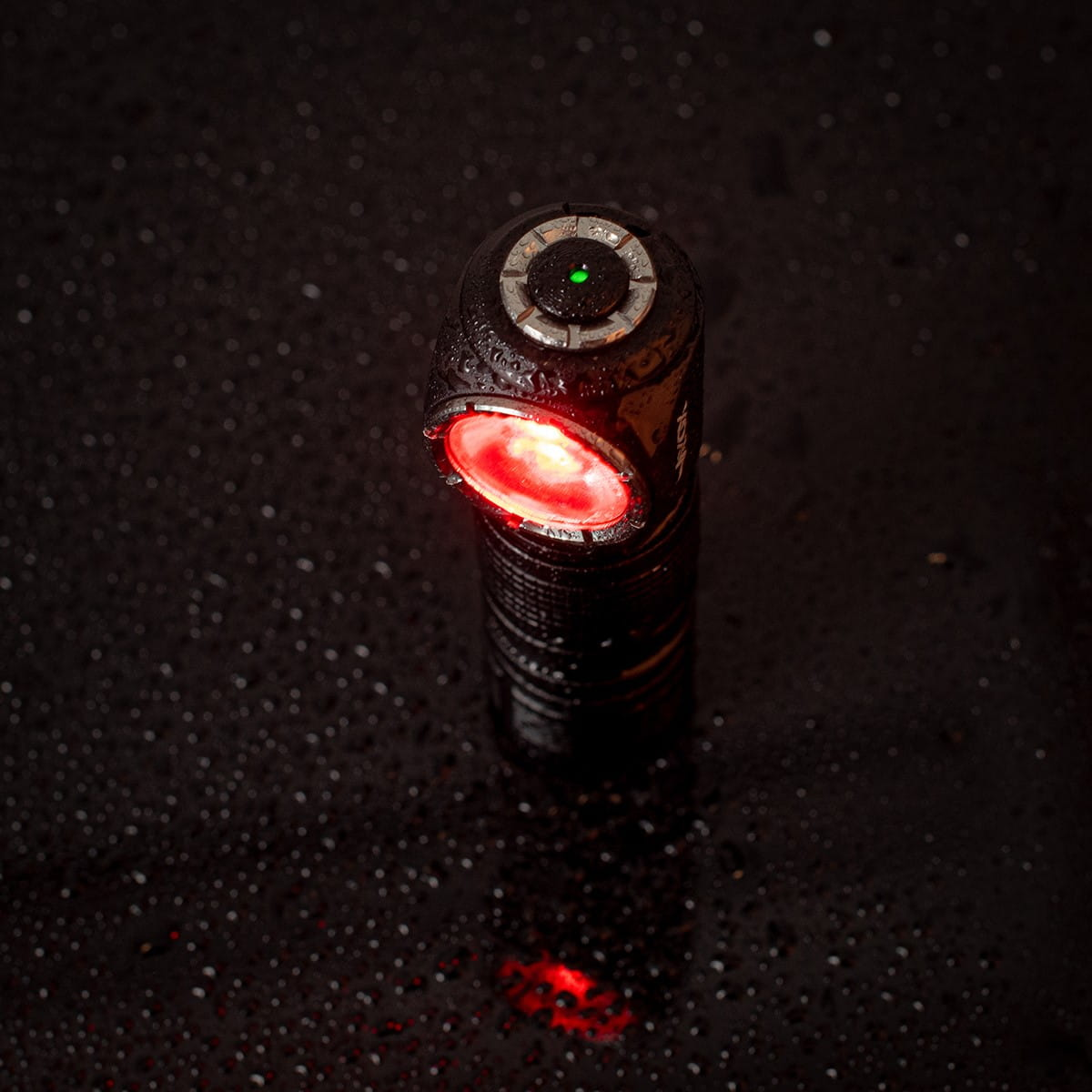 Czarna latarka czołowa z czerwonym światłem na mokrej, ciemnej powierzchni, z kroplami wody odbijającymi światło.