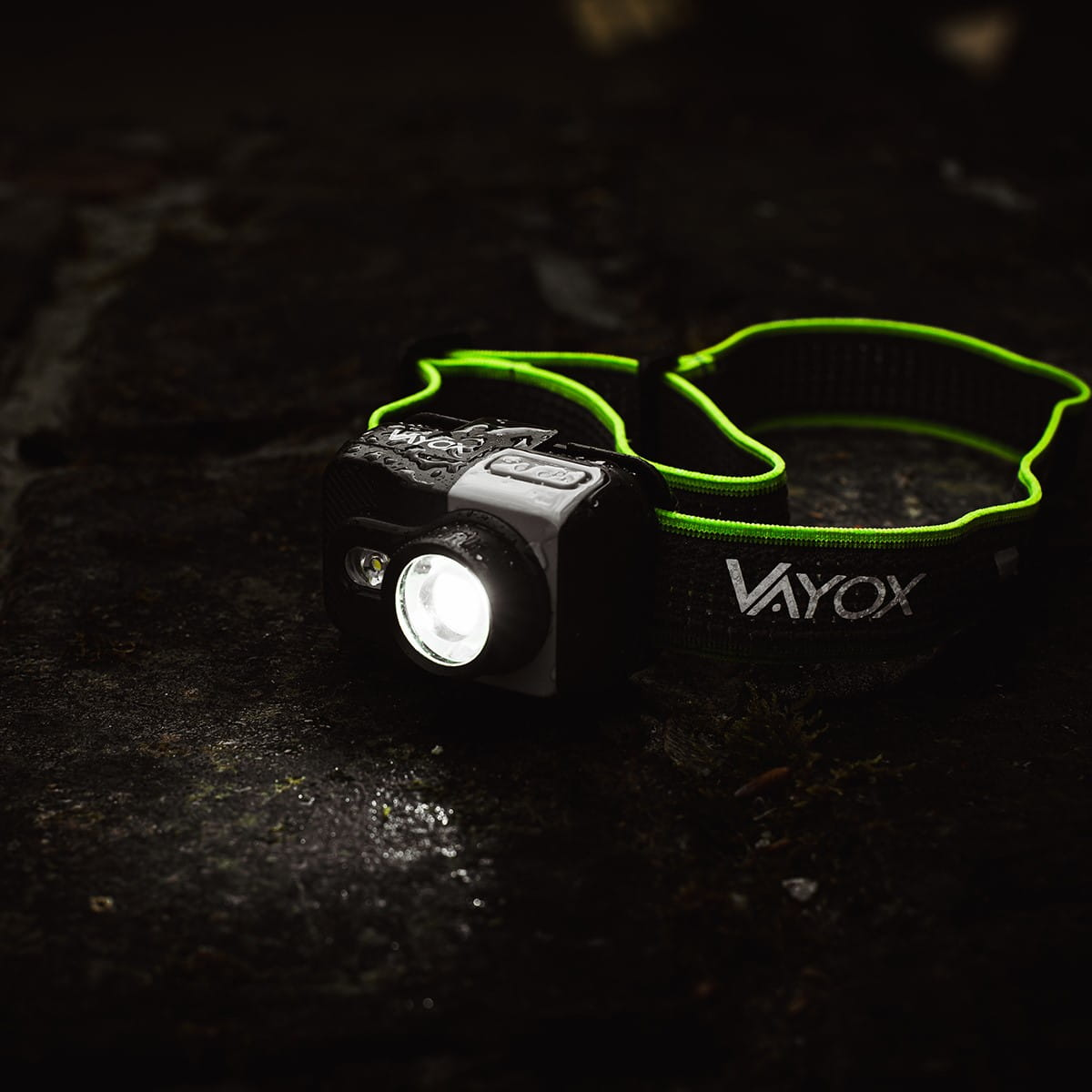 Czarna lampa czołowa z zielonym paskiem, zapalona, leżąca na ciemnej, teksturowanej powierzchni.