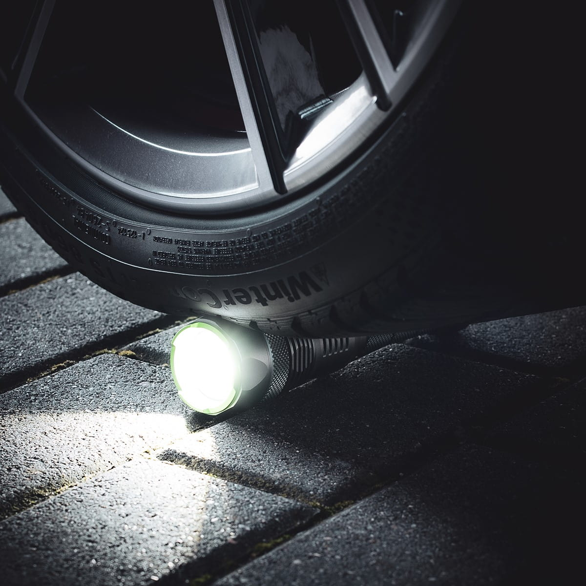 Srebrna latarka z jasnym światłem pod oponą samochodu na ceglanej powierzchni.