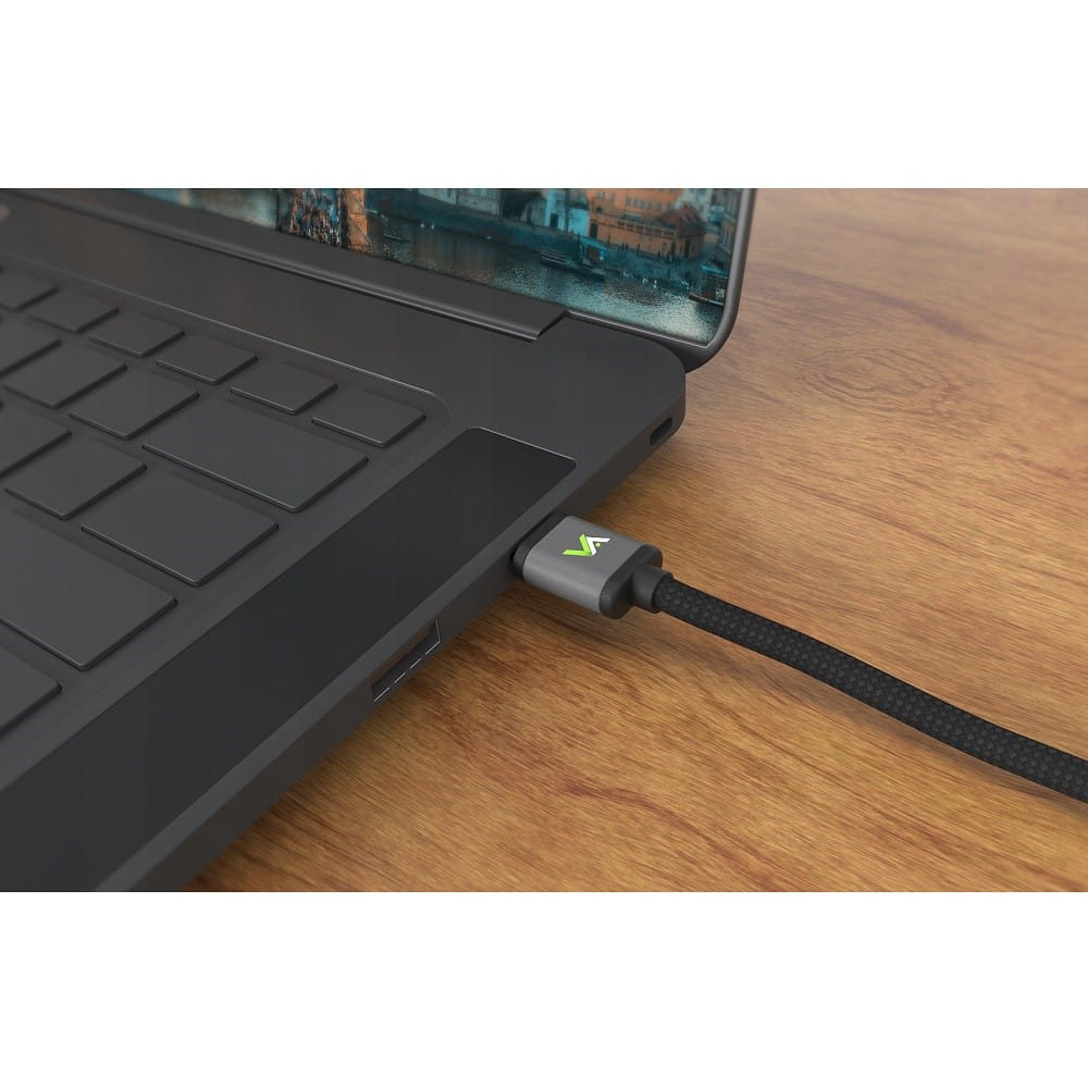 Laptop podłączony czarnym kablem i szarym złączem na drewnianym stole. Ekran pokazuje miasto.