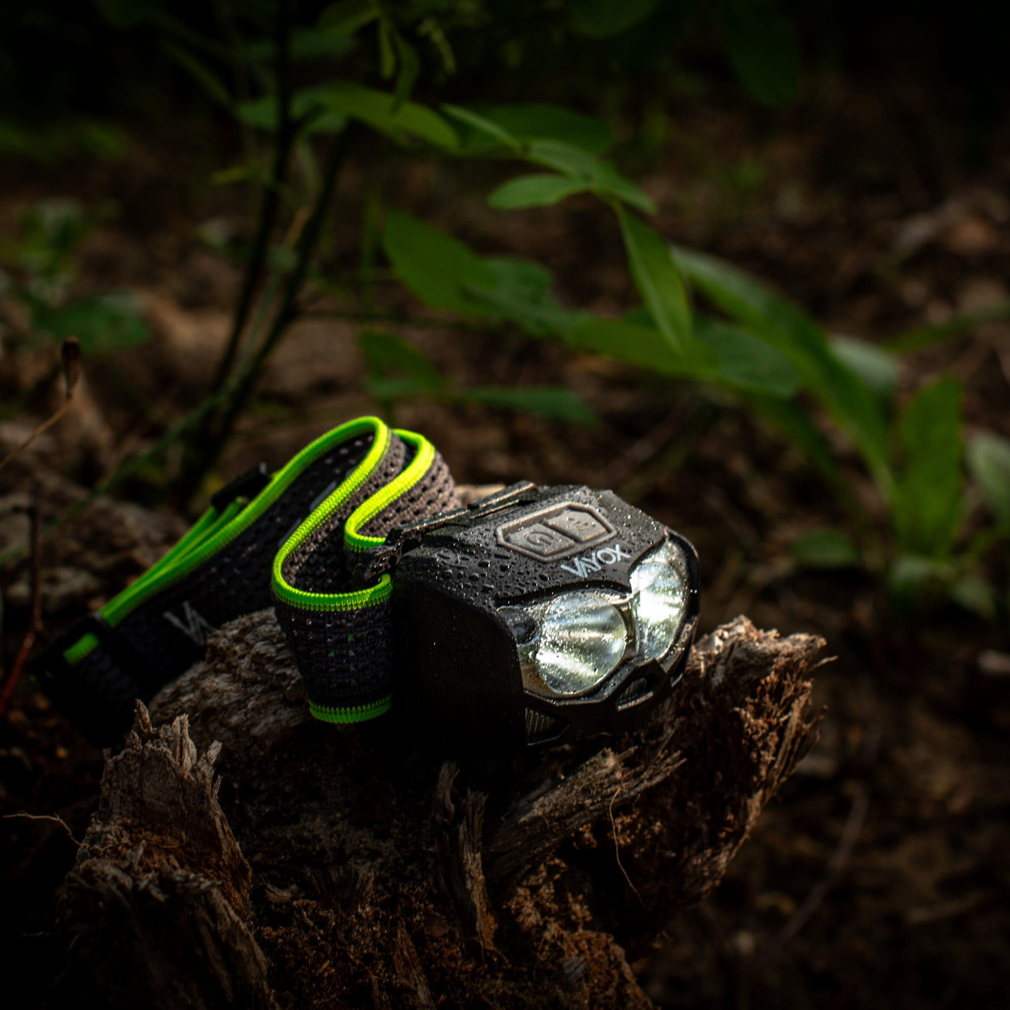 Latarka czołowa na pniu drzewa. Latarka jest czarna z zielonym paskiem. Latarka jest włączona.