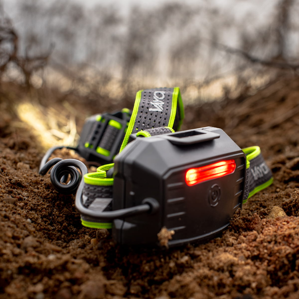 Latarka czołowa na ziemi. Czarna z czerwonym światłem, zielonymi akcentami i logo VAYOX.