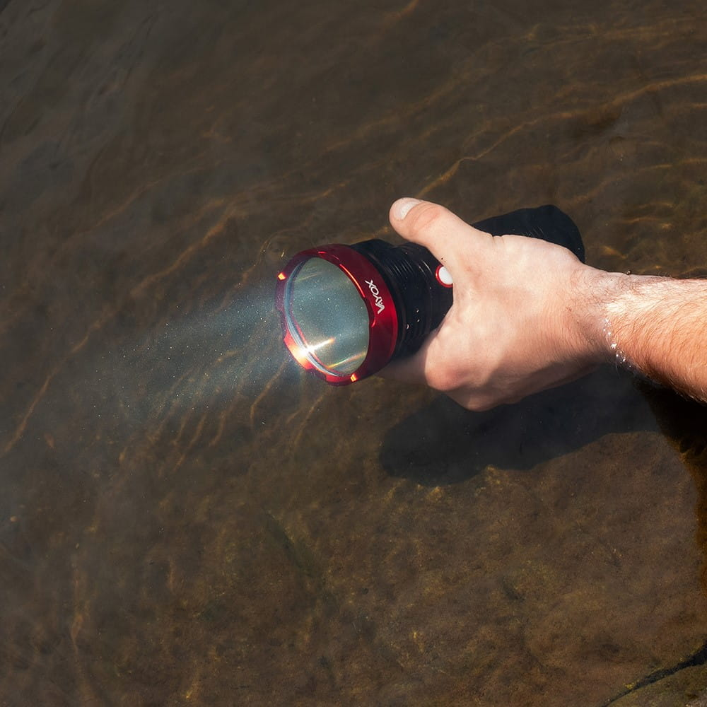 Ręka trzyma czarno-czerwoną latarkę, świecącą w wodzie. Latarka ma czerwony pierścień.