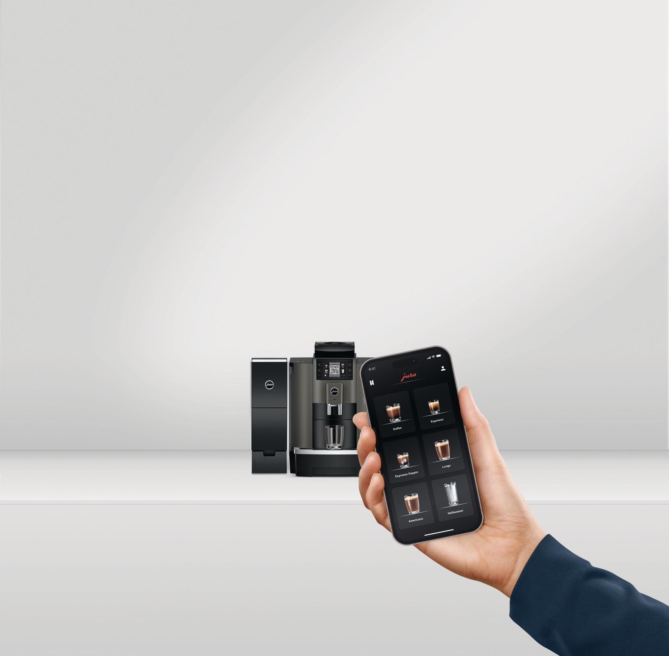 Ręka trzymająca telefon wyświetla aplikację pokazującą różne opcje kawy. Tło: ekspres do kawy.