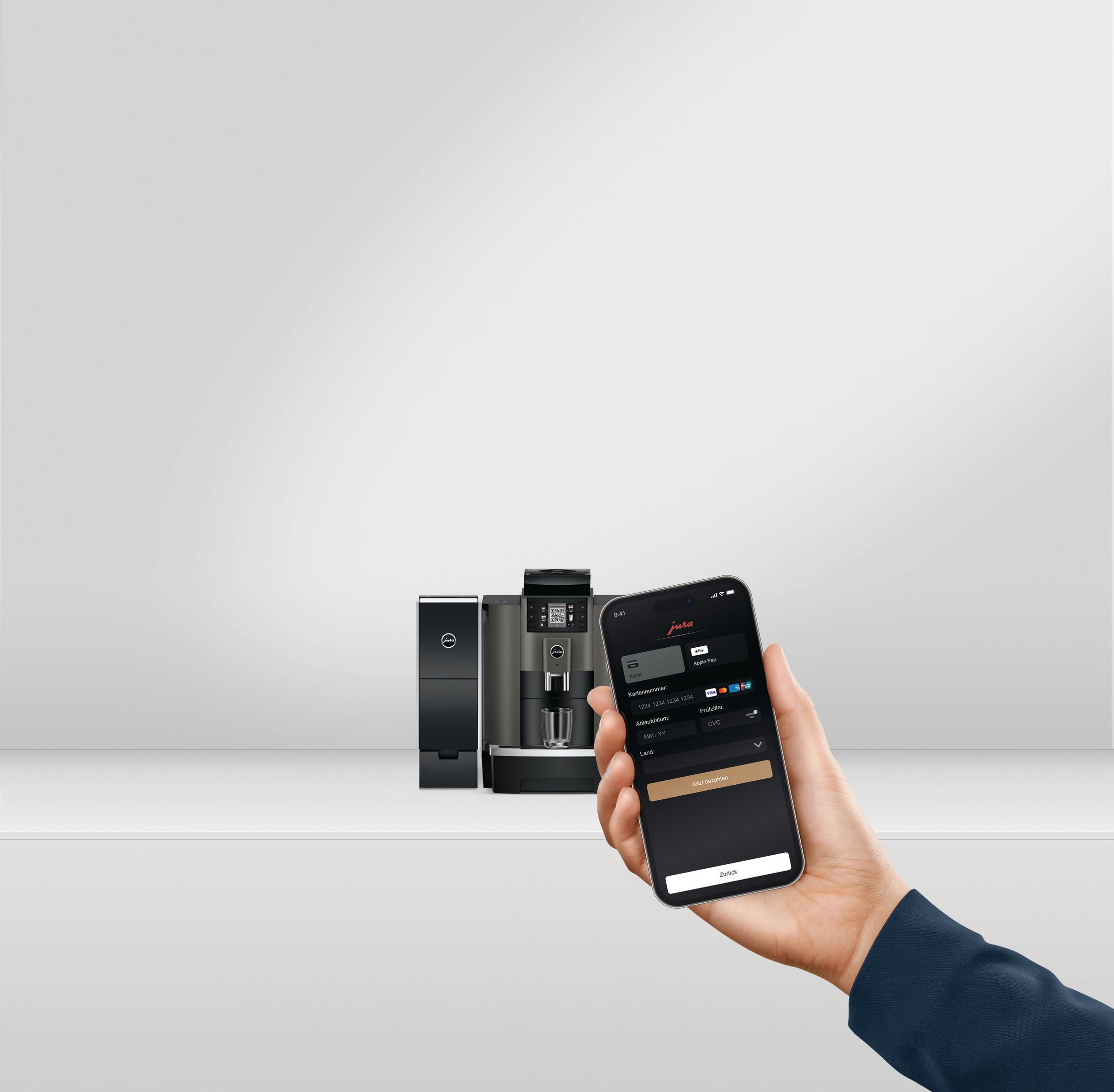 Osoba trzymająca telefon z ekranem kontrolnym ekspresu do kawy. Widoczne ekspresy do kawy.