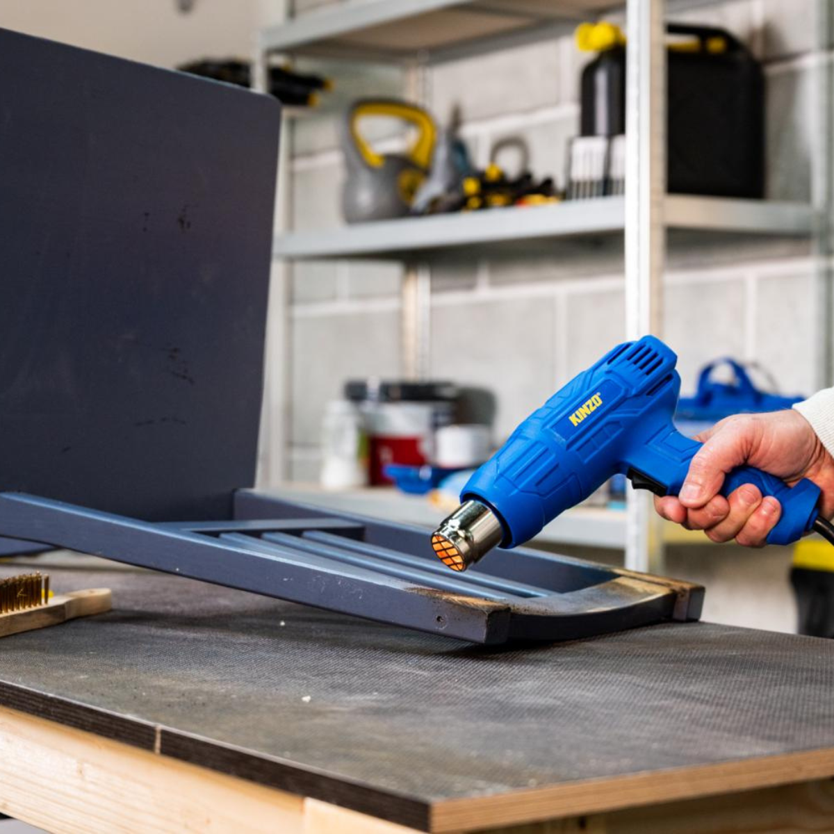 Une personne utilise un pistolet thermique bleu sur une chaise en bois. Le pistolet est dirigé vers le bois. Outils sur une table.