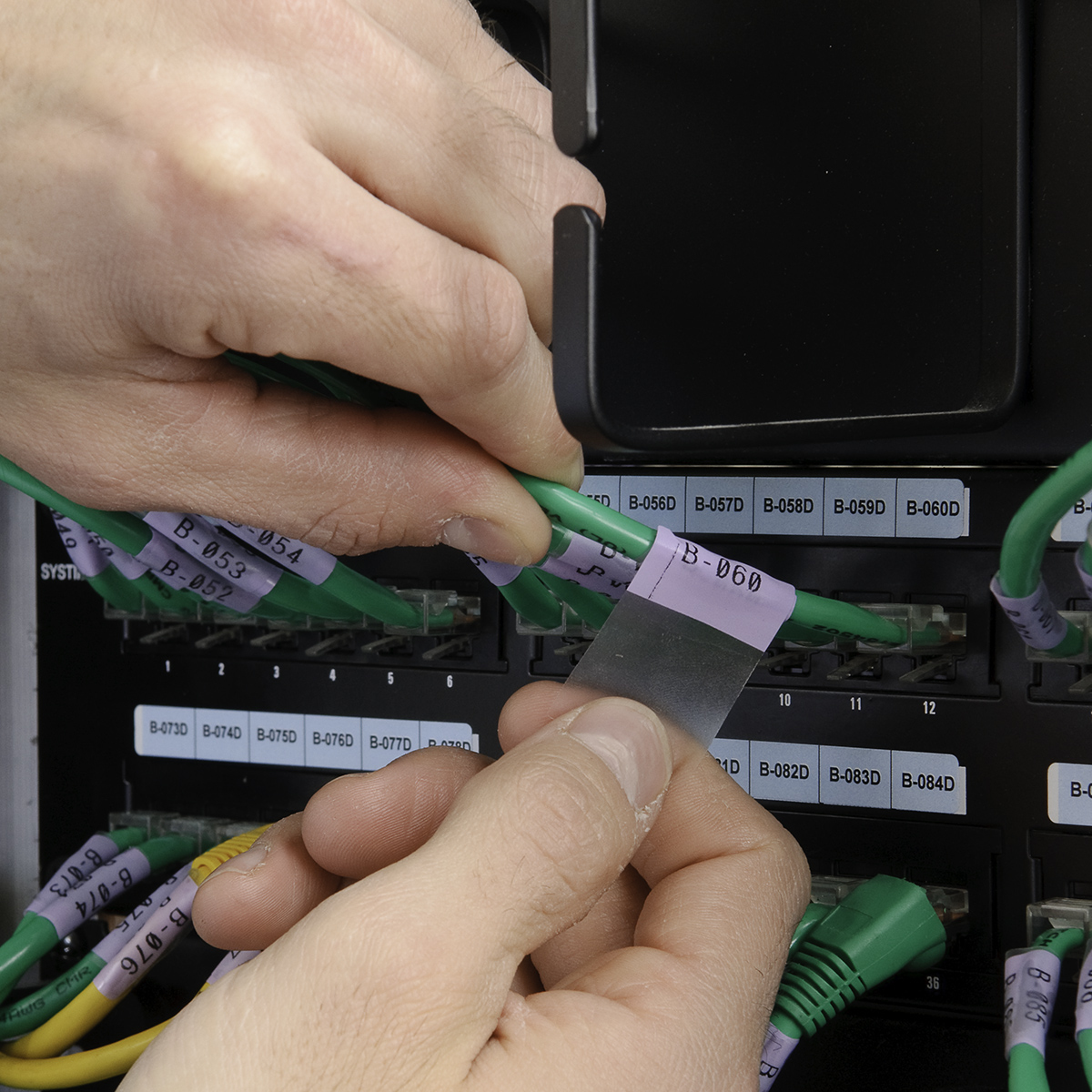Ręce etykietują zielone kable. Kable są podłączone do czarnego panelu. Etykiety mają biały tekst.