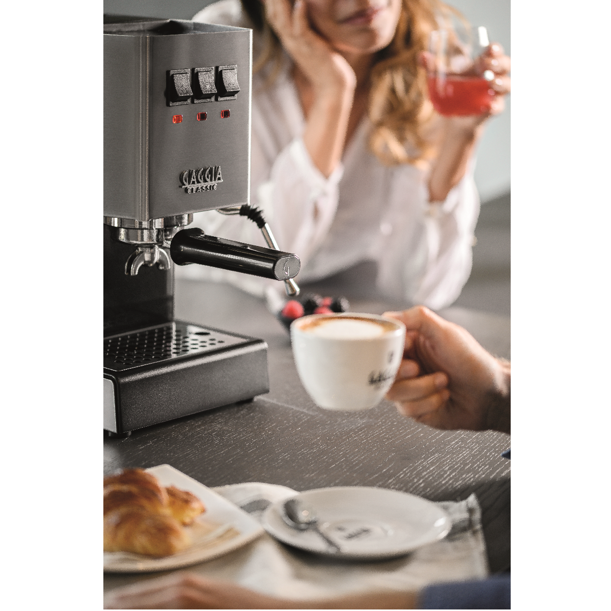 Eine Person hält eine Tasse Kaffee. Eine Kaffeemaschine und ein Croissant liegen auf der Theke.