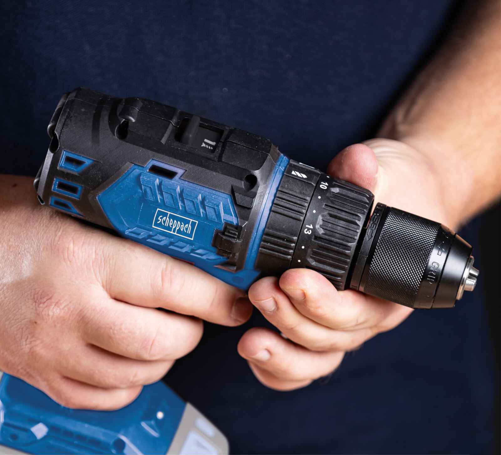 A blue and black cordless drill is held by hands. The drill has a Scheppach logo.