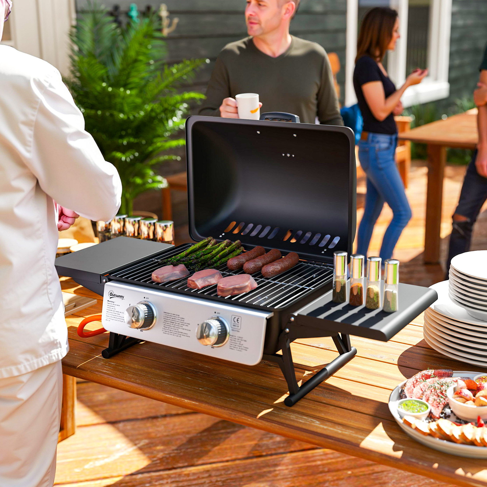 Un barbecue con cibo su un tavolo di legno, circondato da persone, cibo e piatti.