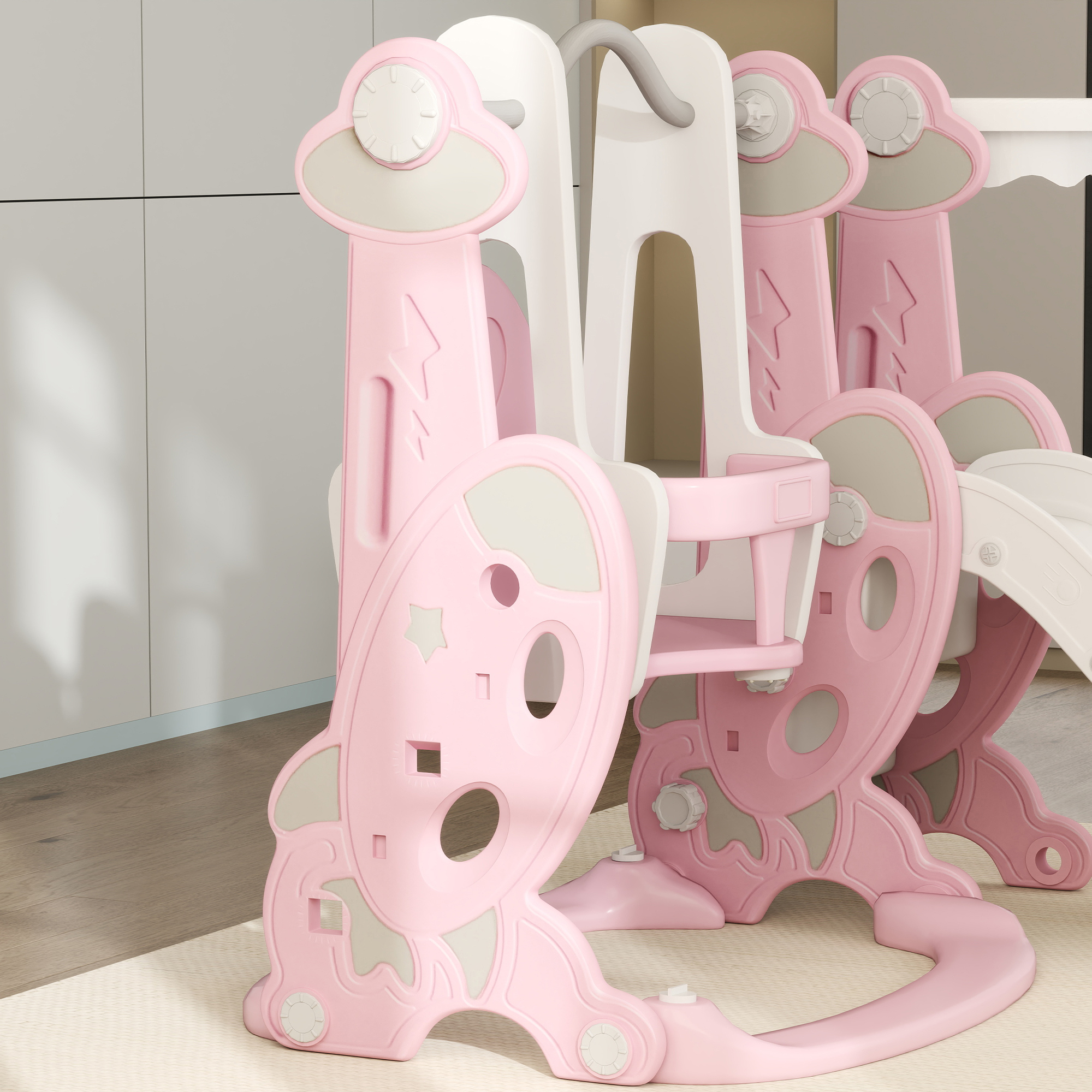 Un columpio para bebés en forma de cohete rosa y blanco con un asa y detalles grises, sobre una alfombra.
