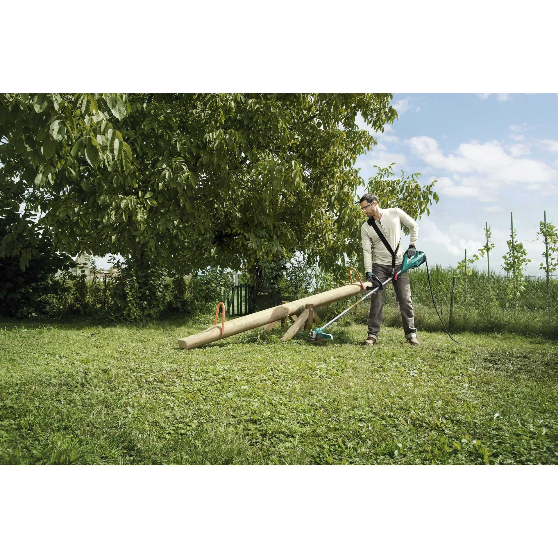 Ein Mann benutzt einen Trimmer auf einem Rasen mit einer Wippe im Hintergrund.