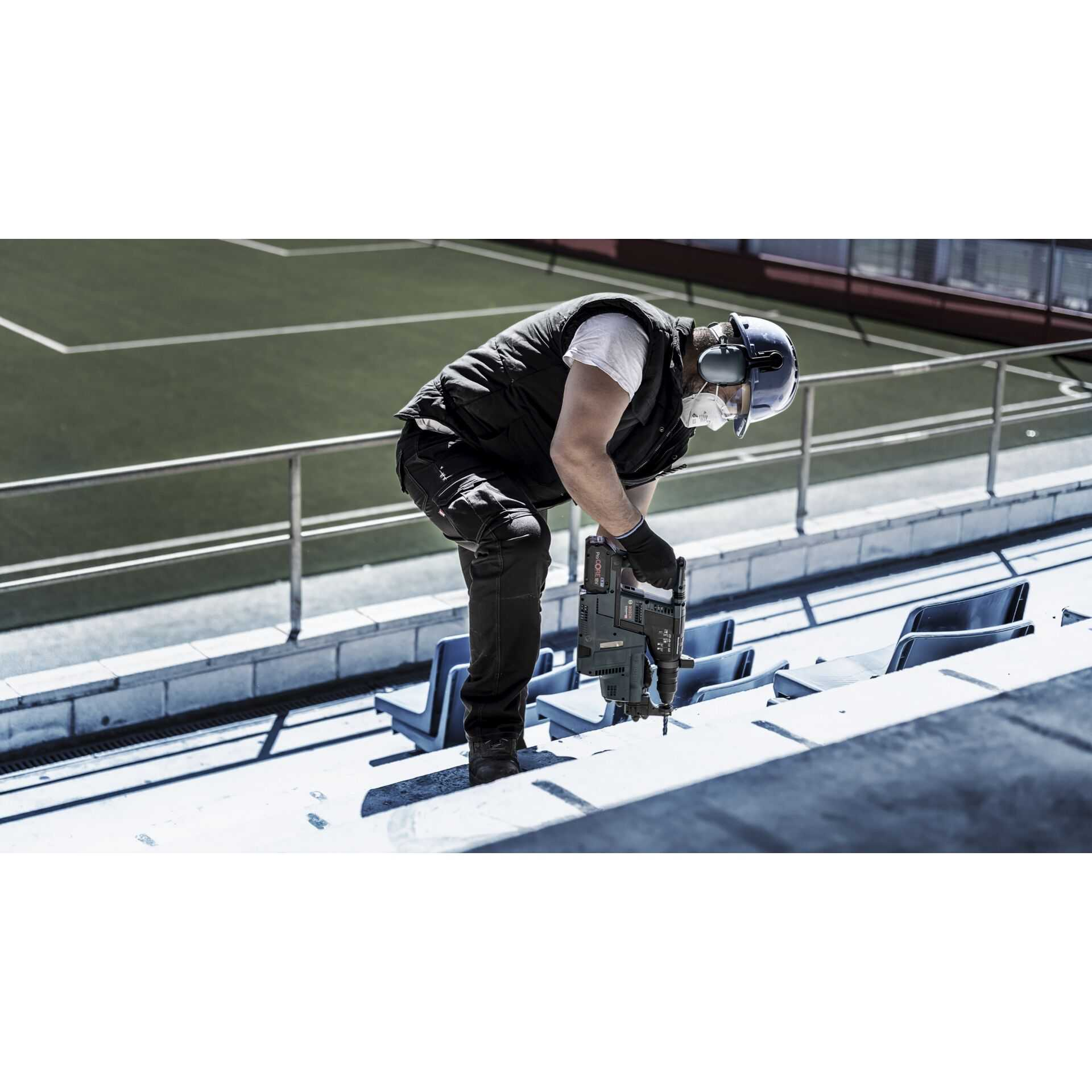 Osoba wierci w betonowych stopniach trybuny. Noszą sprzęt ochronny. Używana jest wiertarka.